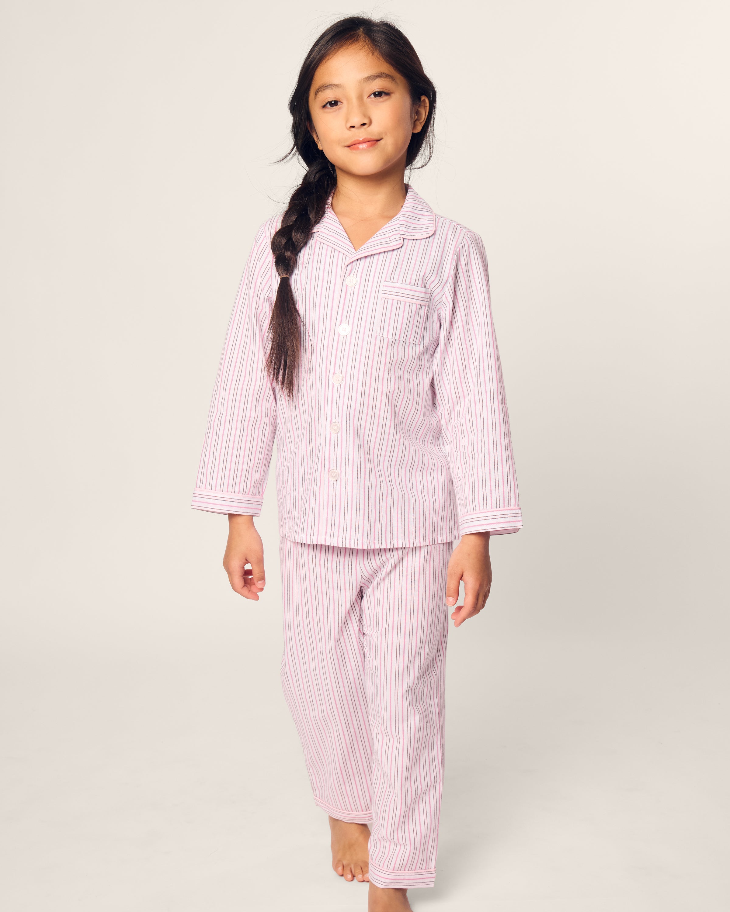 A young child with a long dark braid stands barefoot against a plain background, wearing the Petite Plume Kids Twill Pajama in Blush Boulevard.