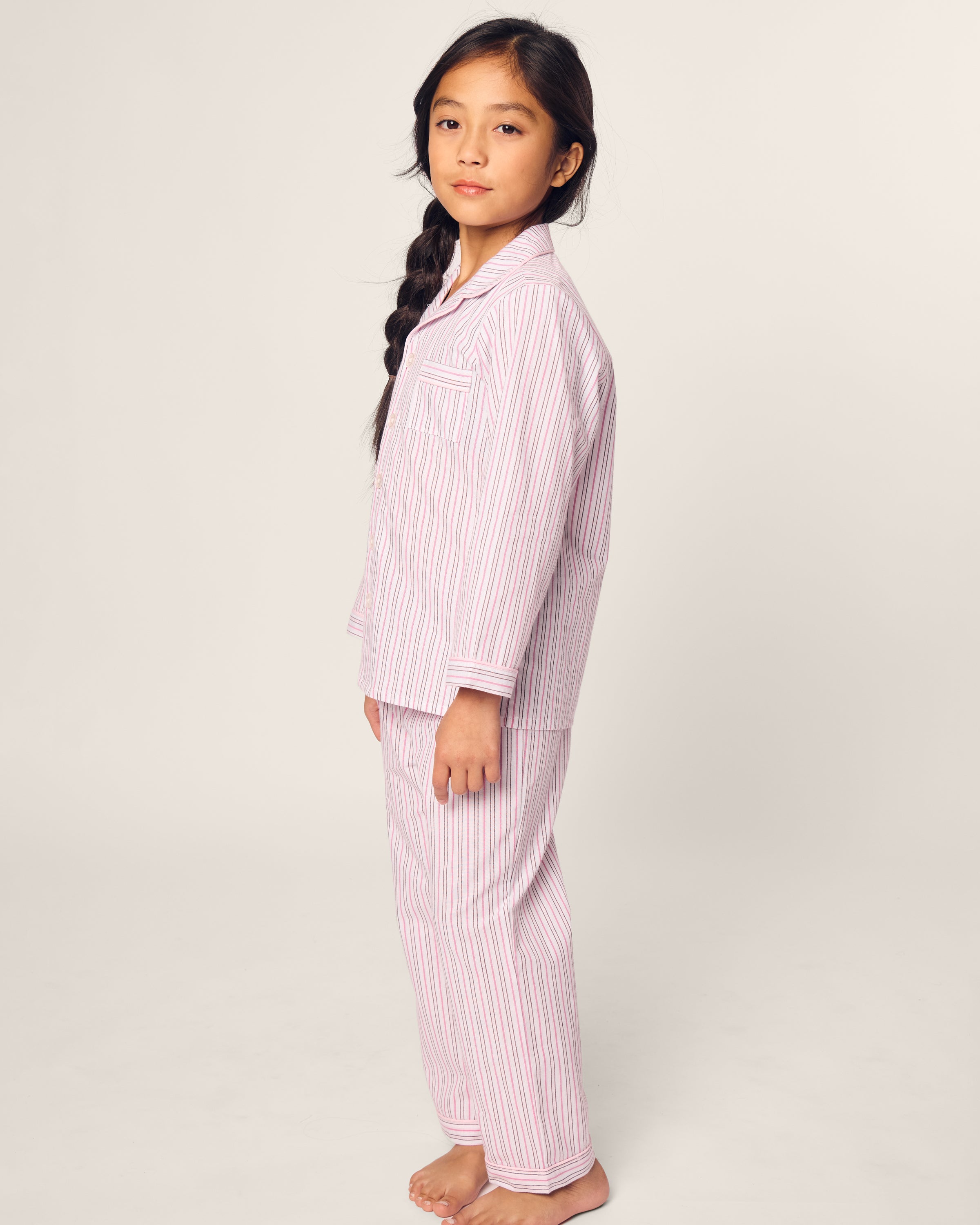 A young girl, with long braided hair and bare feet, stands sideways in Petite Plumes Kids Twill Pajama in Blush Boulevard. This pink and white striped set, made from soft yarn-dyed cotton, catches the cameras gaze against a plain light background.