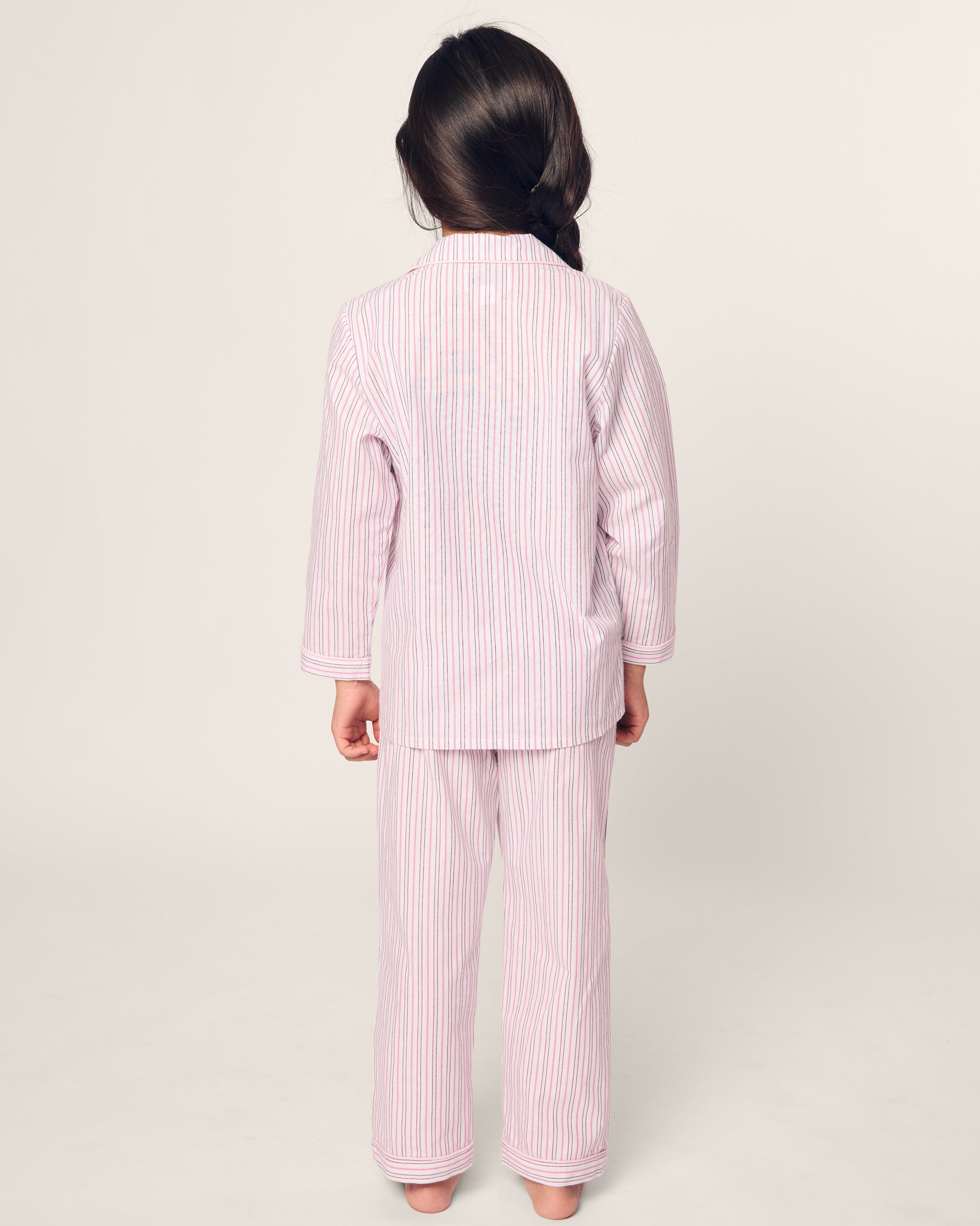 A child wears Petite Plumes Kids Twill Pajama in Blush Boulevard, featuring pink and white stripes. They stand barefoot with their back to the camera, while a long braid hangs against a plain light backdrop.