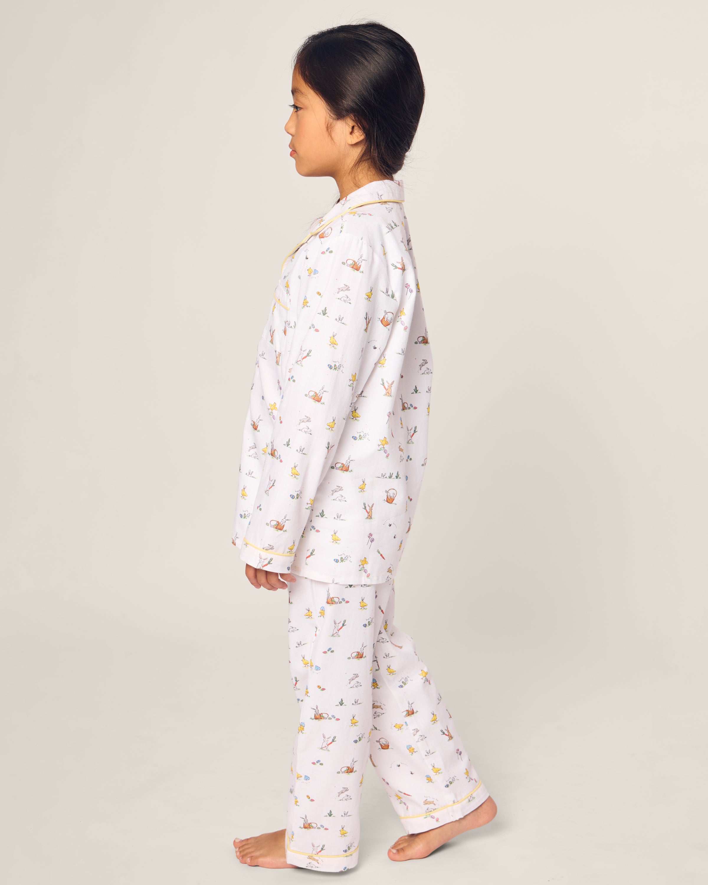 A young girl stands barefoot in profile against a plain background, wearing Petite Plumes Kids Twill Pajama in Easter Frolic, featuring a small print pattern. Her hair is styled in a neat bun, exuding an air of timeless charm.