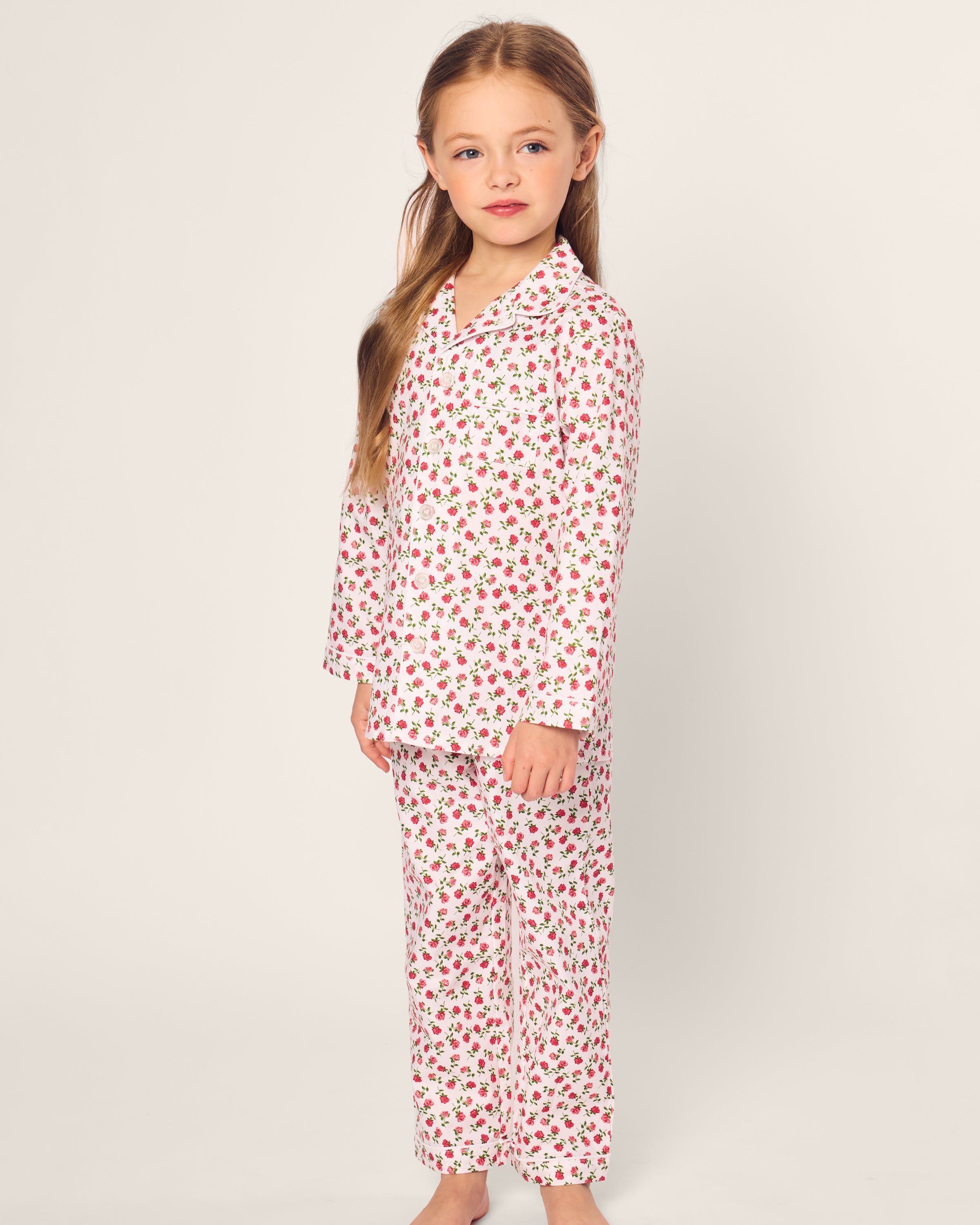 A young girl with long light brown hair stands barefoot against a neutral background, wearing Petite Plumes Kids Twill Pajama in Petite Petals, made from yarn-dyed cotton sleepwear. She looks straight ahead with a slight smile.