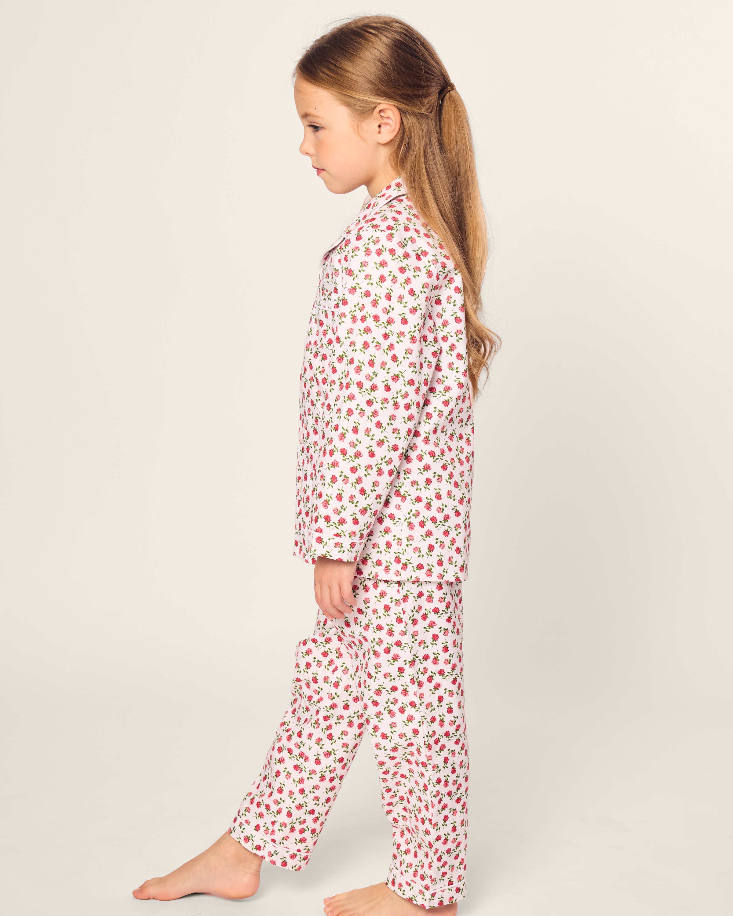 A young girl with long hair stands barefoot on a plain background, facing sideways, wearing Petite Plumes Kids Twill Pajama in Petite Petals.