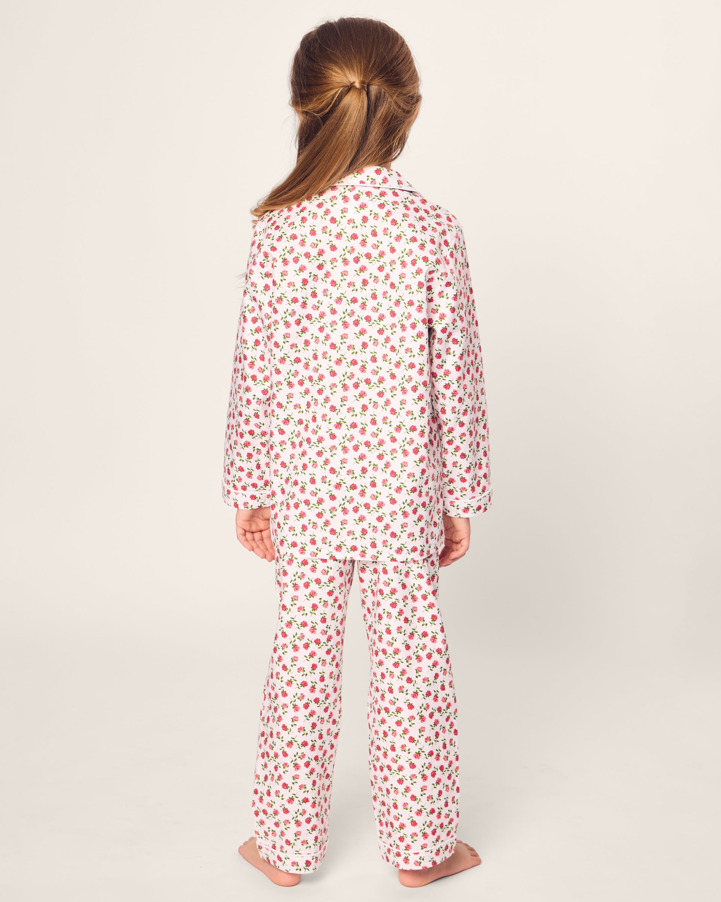 A child in Petite Plume’s Kids Twill Pajama in Petite Petals stands barefoot on a plain background, their long brown hair accentuated by pink flowers against the matching fabric.