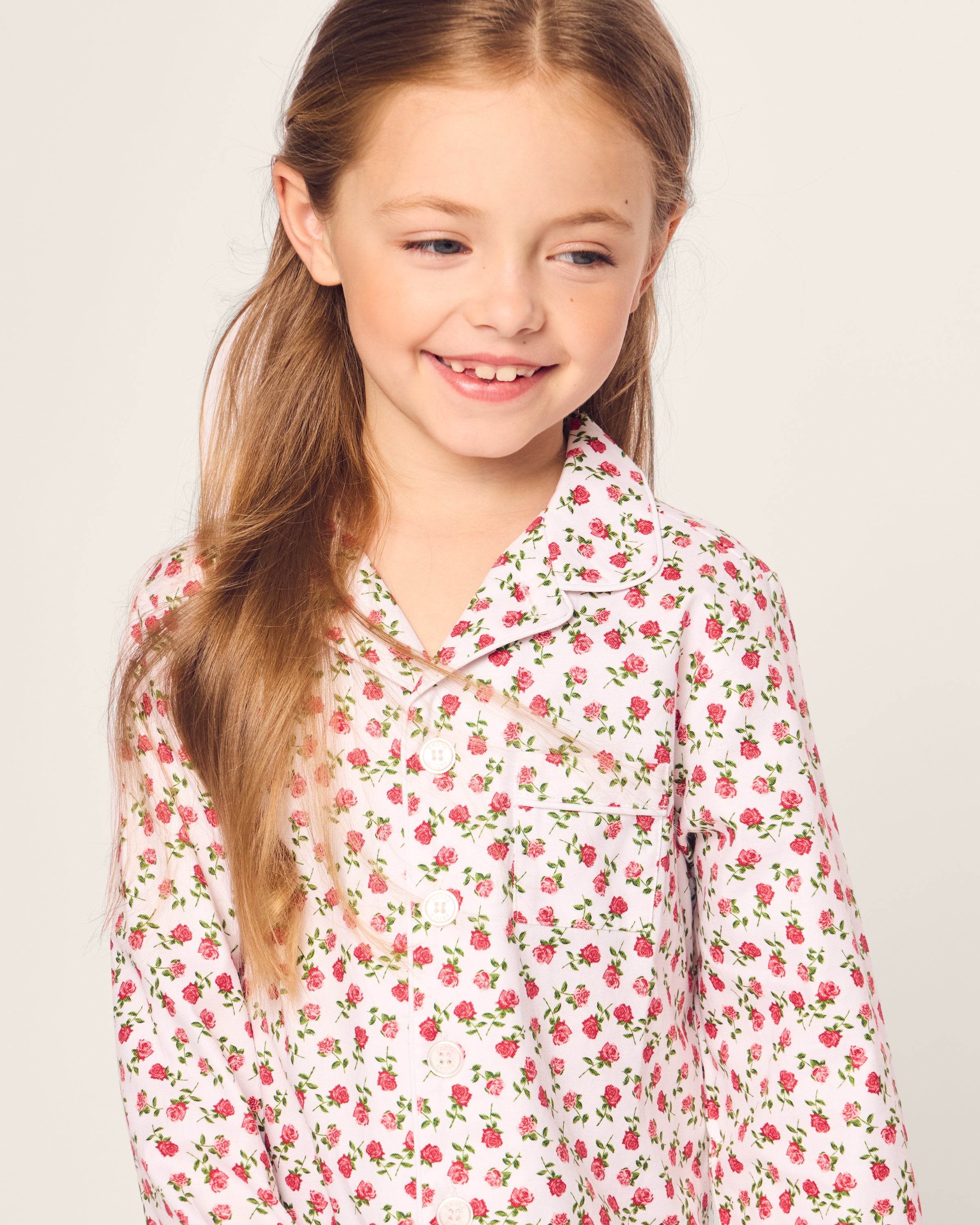 A young girl with long hair, smiling and gazing to the side, wears a Petite Plume Kids Twill Pajama in Petite Petals against a plain background.