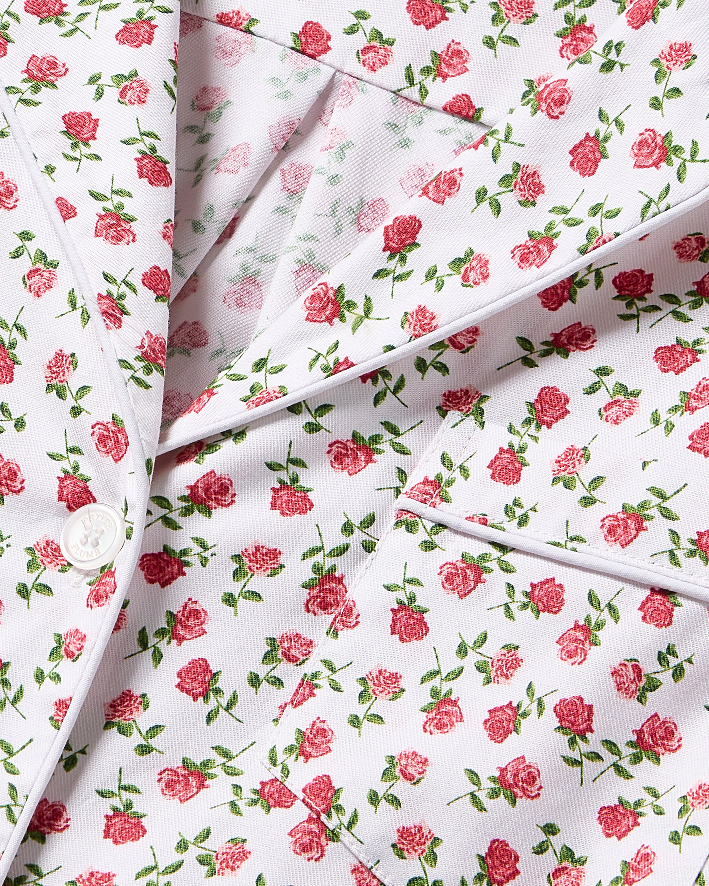 Close-up of the Kids Twill Pajama in Petite Petals by Petite Plume, featuring a white fabric with red and green rose patterns. Designed in a suit jacket style with buttons, lapels, and pockets, the floral pattern covers the entire surface.