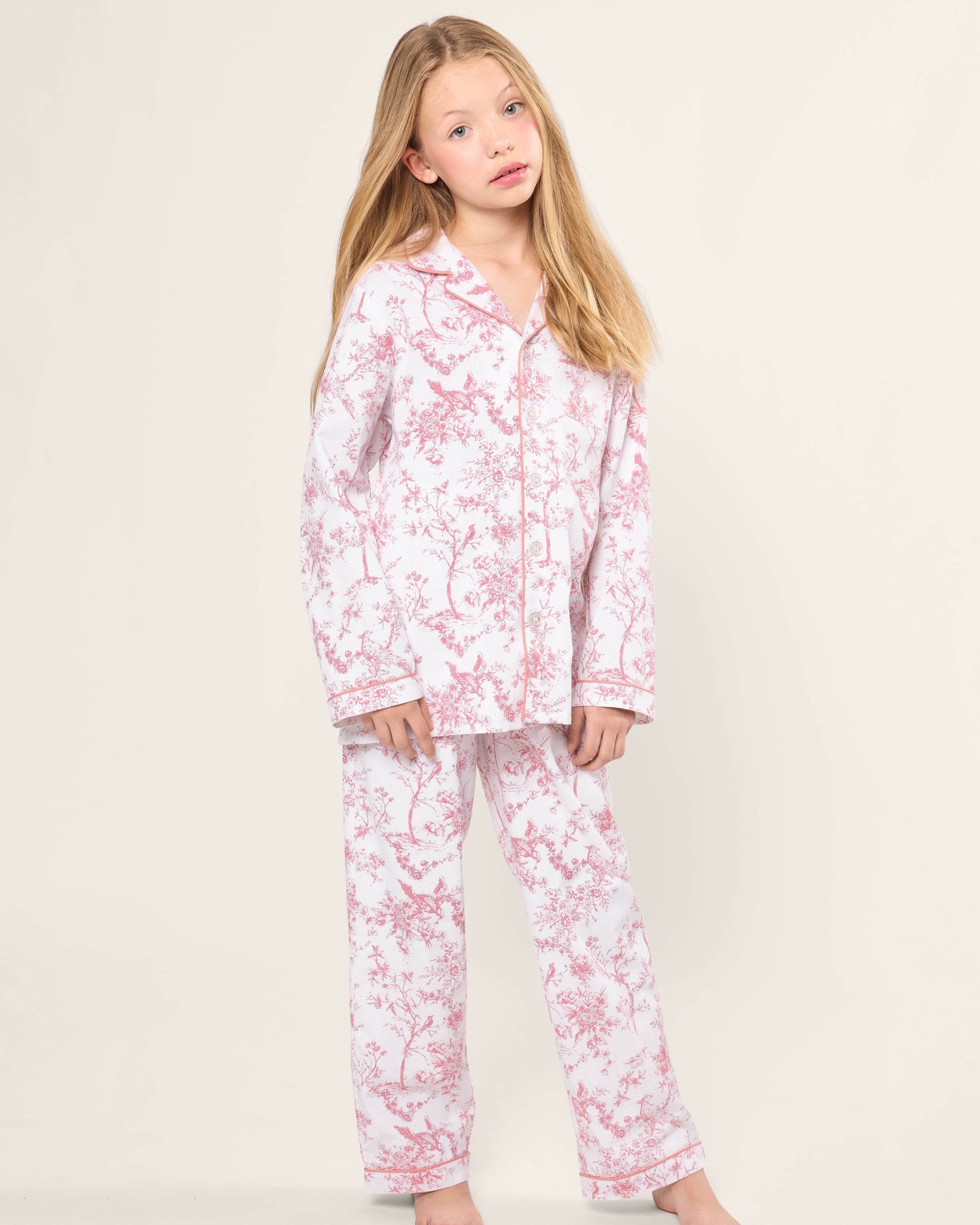 A young girl with long blonde hair stands barefoot against a plain light background, wearing a luxurious Pajama Set by Petite Plume. The Kids Twill Pajama Set in Pink Timeless Toile complements her serene gaze looking slightly off-camera with its botanical elegance of yarn-dyed cotton fabric.