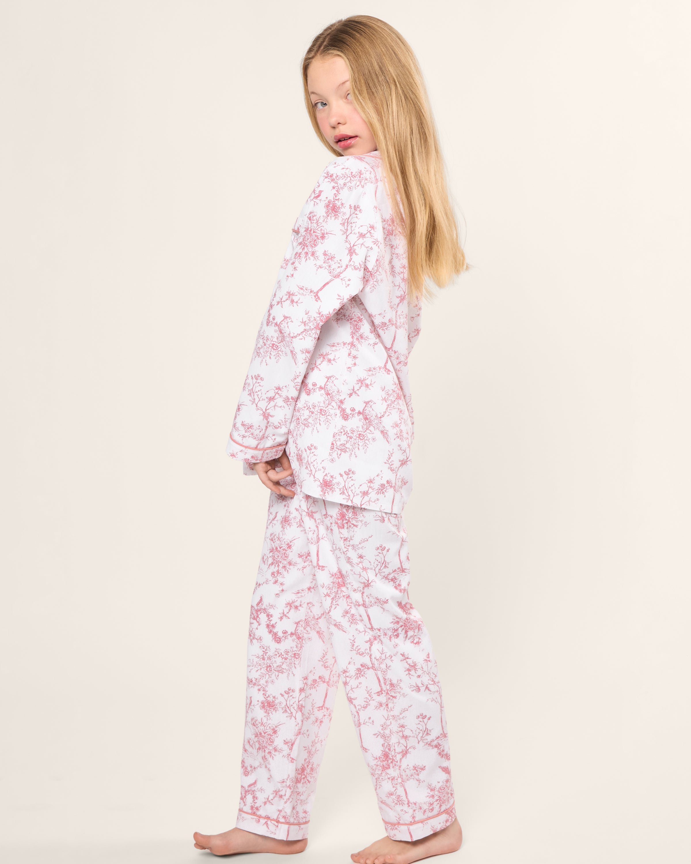 A child with long blonde hair poses barefoot against a plain backdrop, wearing Petite Plumes Kids Twill Pajama Set in Pink Timeless Toile. The pajama set features pink and white floral patterns, and the child glances towards the camera, embodying botanical elegance.