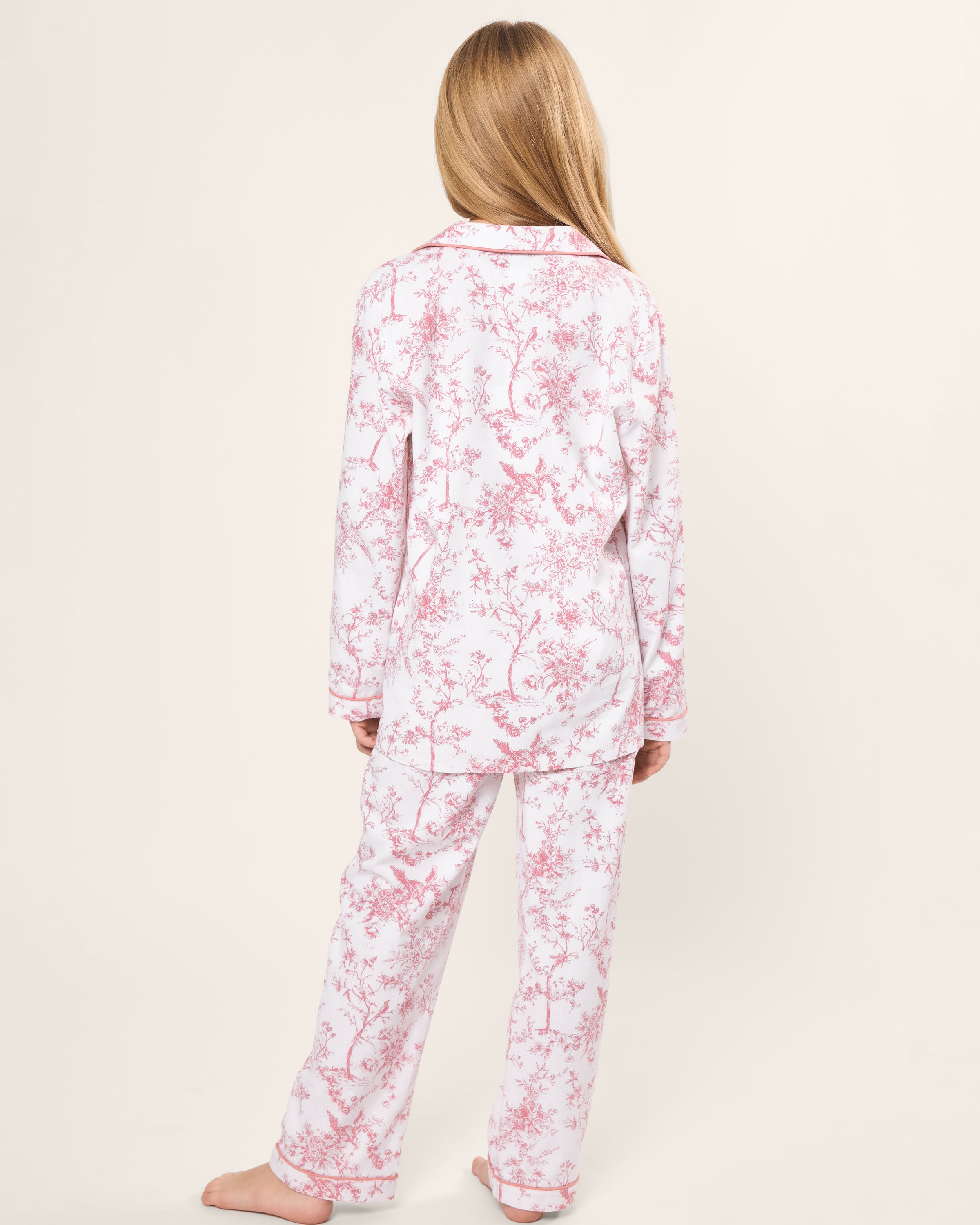 A person with long hair wears the Kids Twill Pajama Set in Pink Timeless Toile by Petite Plume, crafted from yarn-dyed cotton with a pink floral design on a white background. They stand barefoot, exuding botanical elegance facing away from the camera.