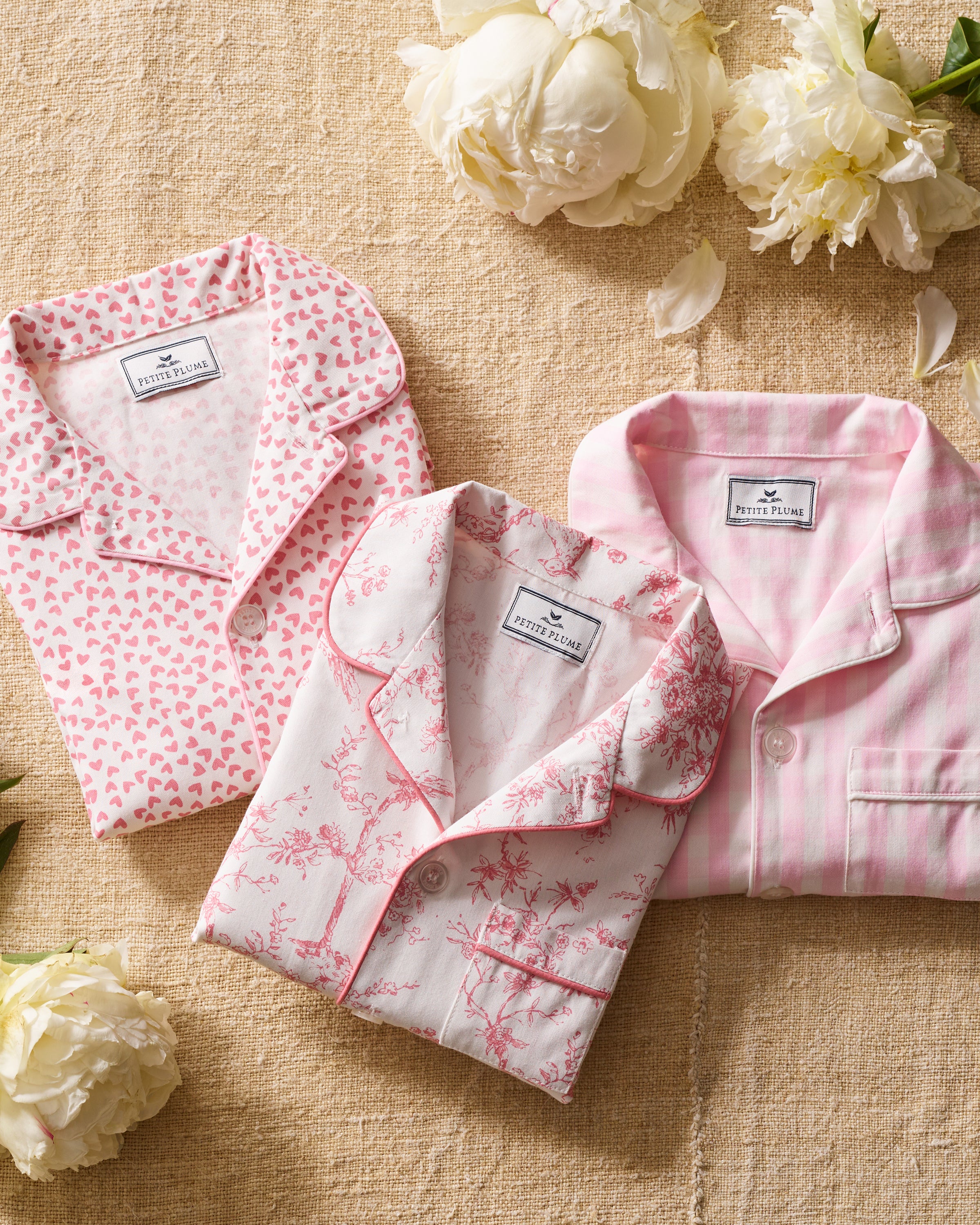 Three folded Petit Plume Kids Twill Pajama tops, styled in luxury sleepwear, are arranged on a beige fabric. They boast patterns of pink hearts, pink-and-white toile, and pink stripes. Scattered white peonies and petals add a touch of botanical elegance.