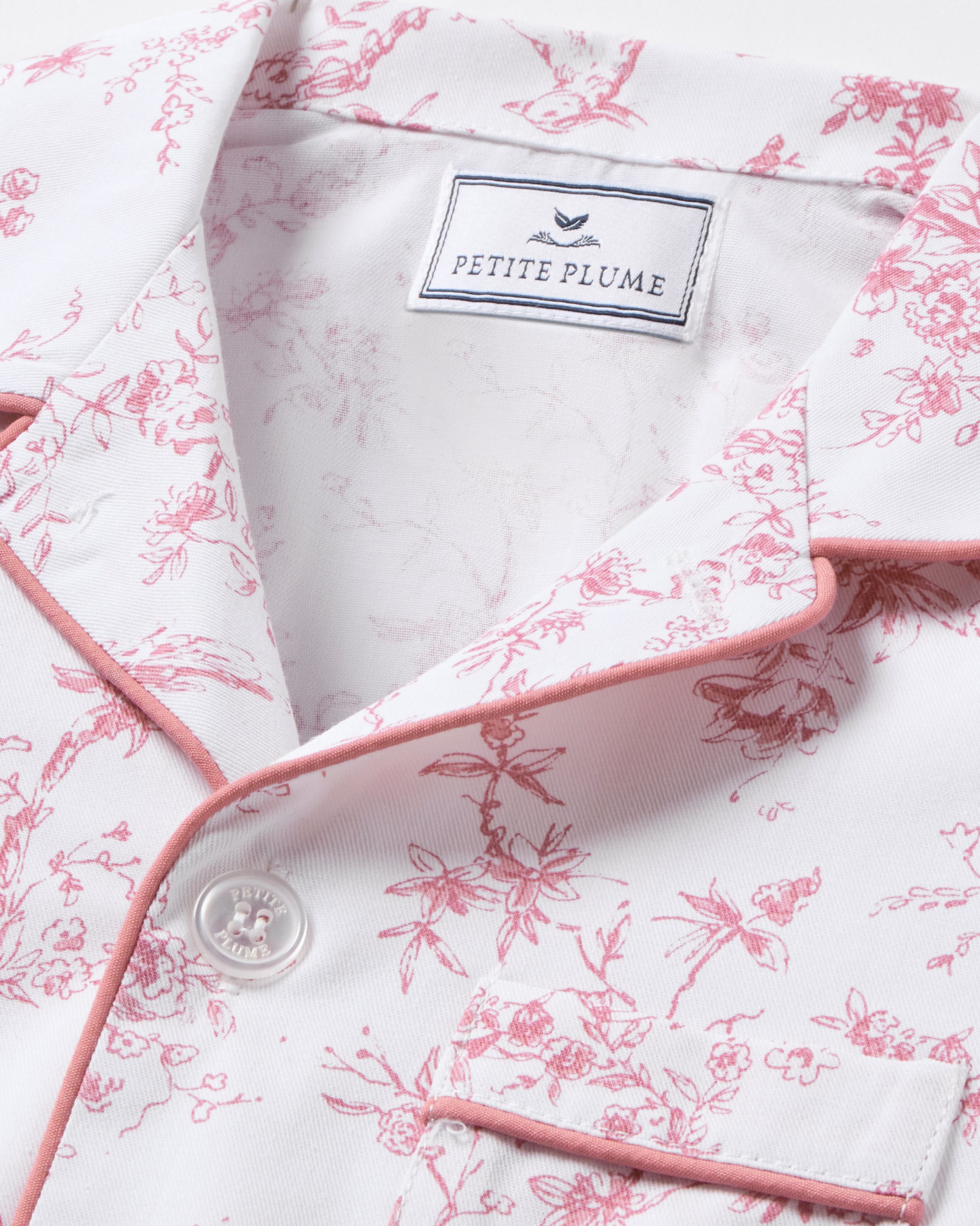 A close-up of Petite Plumes Kids Twill Pajama Set in Pink Timeless Toile shows a white top with pink floral patterns and piping. It features a button-up front, a small pocket, and the Petite Plume label with a bird logo, epitomizing botanical elegance in luxury sleepwear.