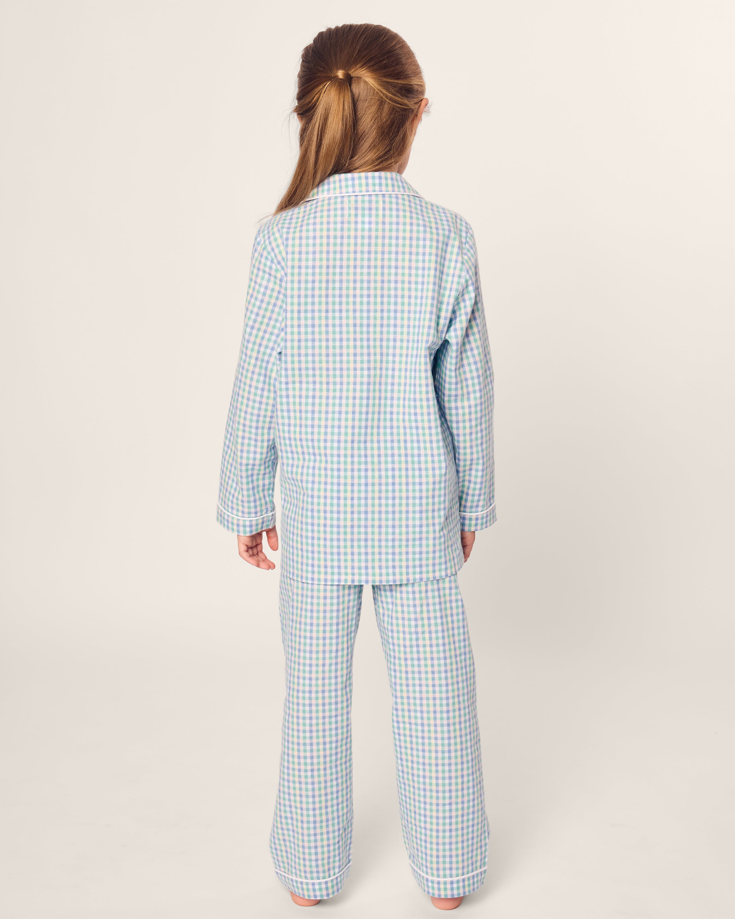 A child with long, light brown hair faces away, wearing a Petite Plume Kids Twill Pajama in Spring Gingham. The blue and white checkered set is crafted from yarn-dyed cotton with a flame retardant design for safety against a plain, light-colored background.