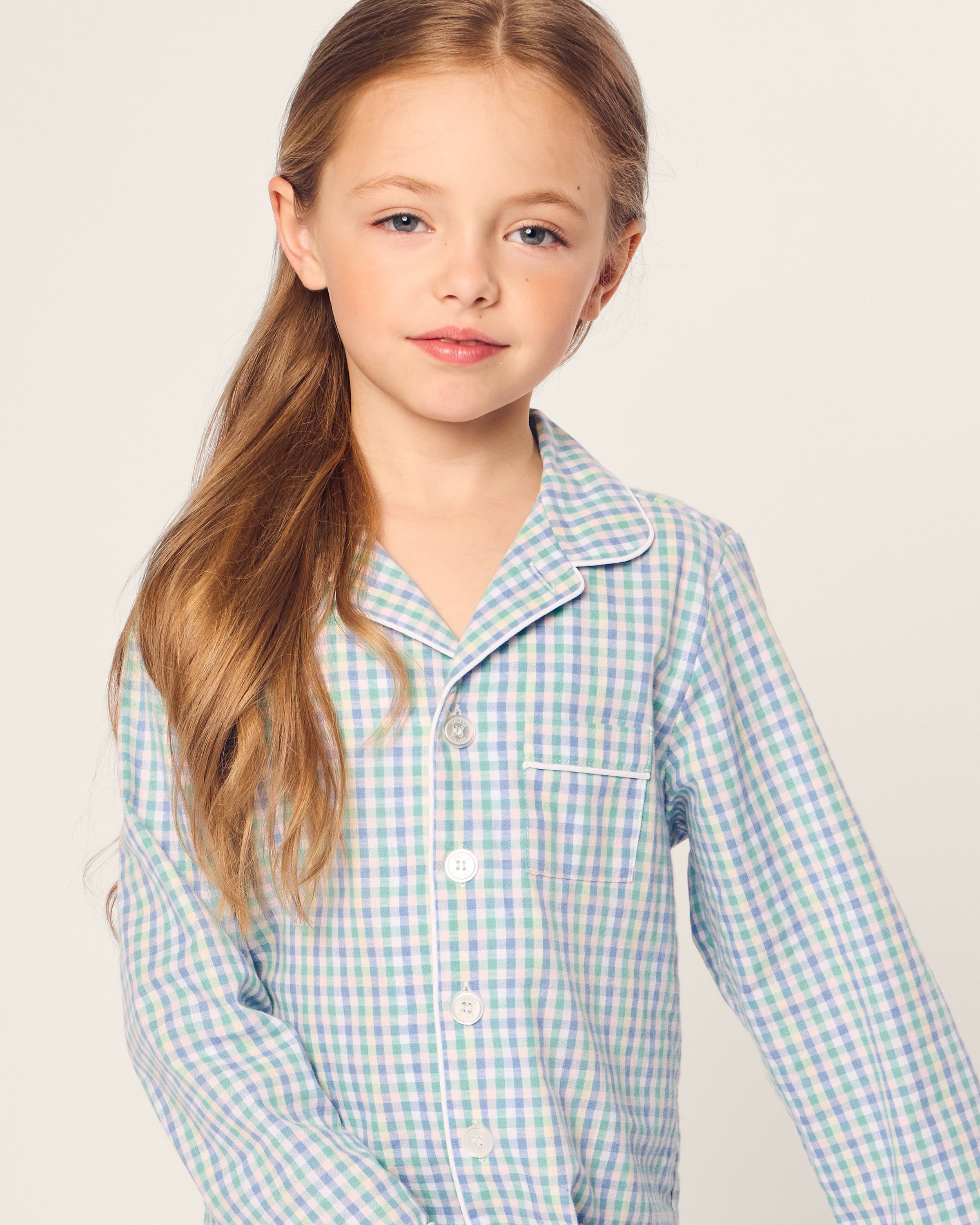 A young girl with long brown hair wears Petite Plumes Kids Twill Pajama in Spring Gingham. She poses against a plain, light background, looking calmly at the camera.