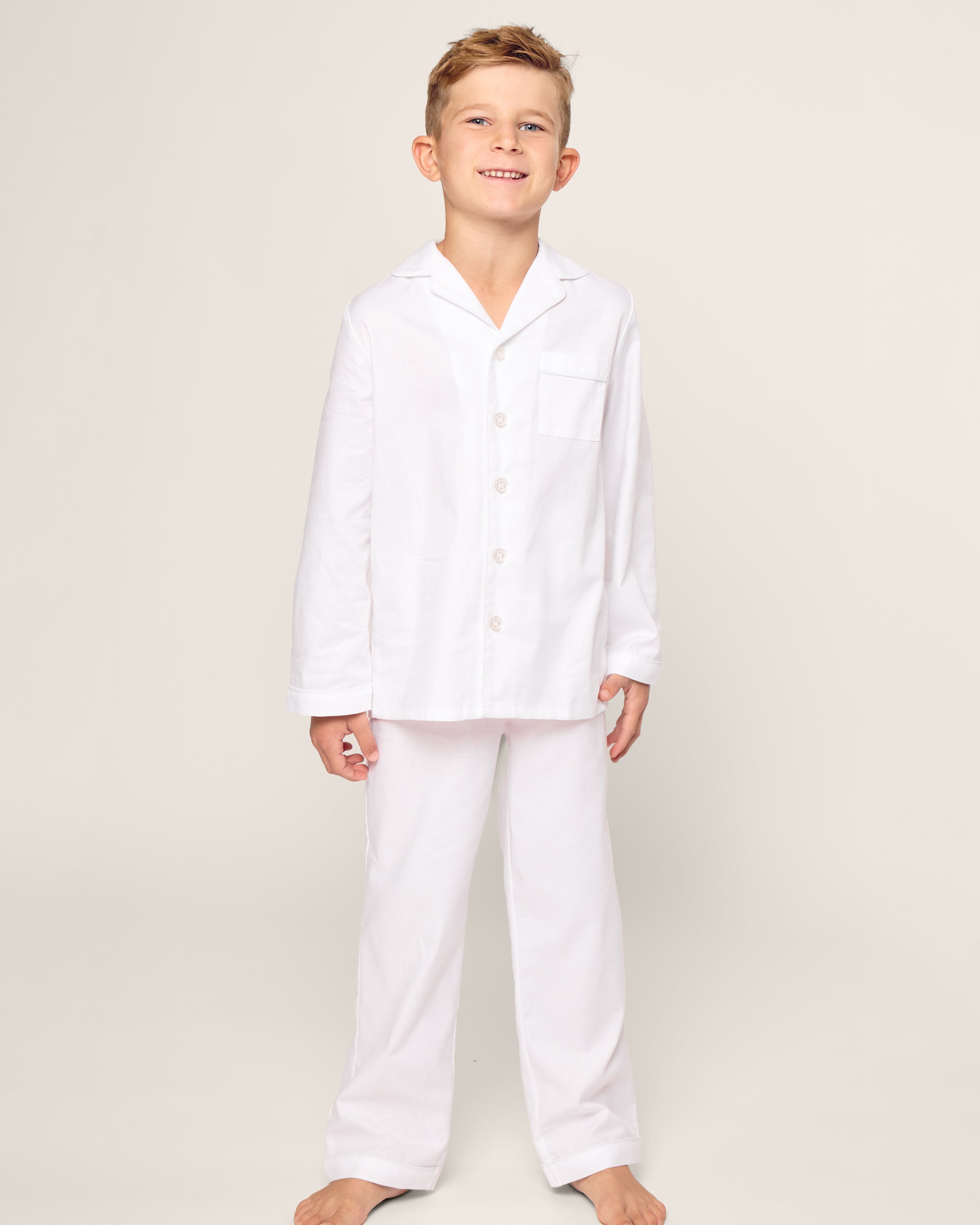 A smiling child stands barefoot in a luxurious Petite Plume sleepwear set, wearing the Kids Twill Pajama Set in White with White Piping, against a plain light-colored background.