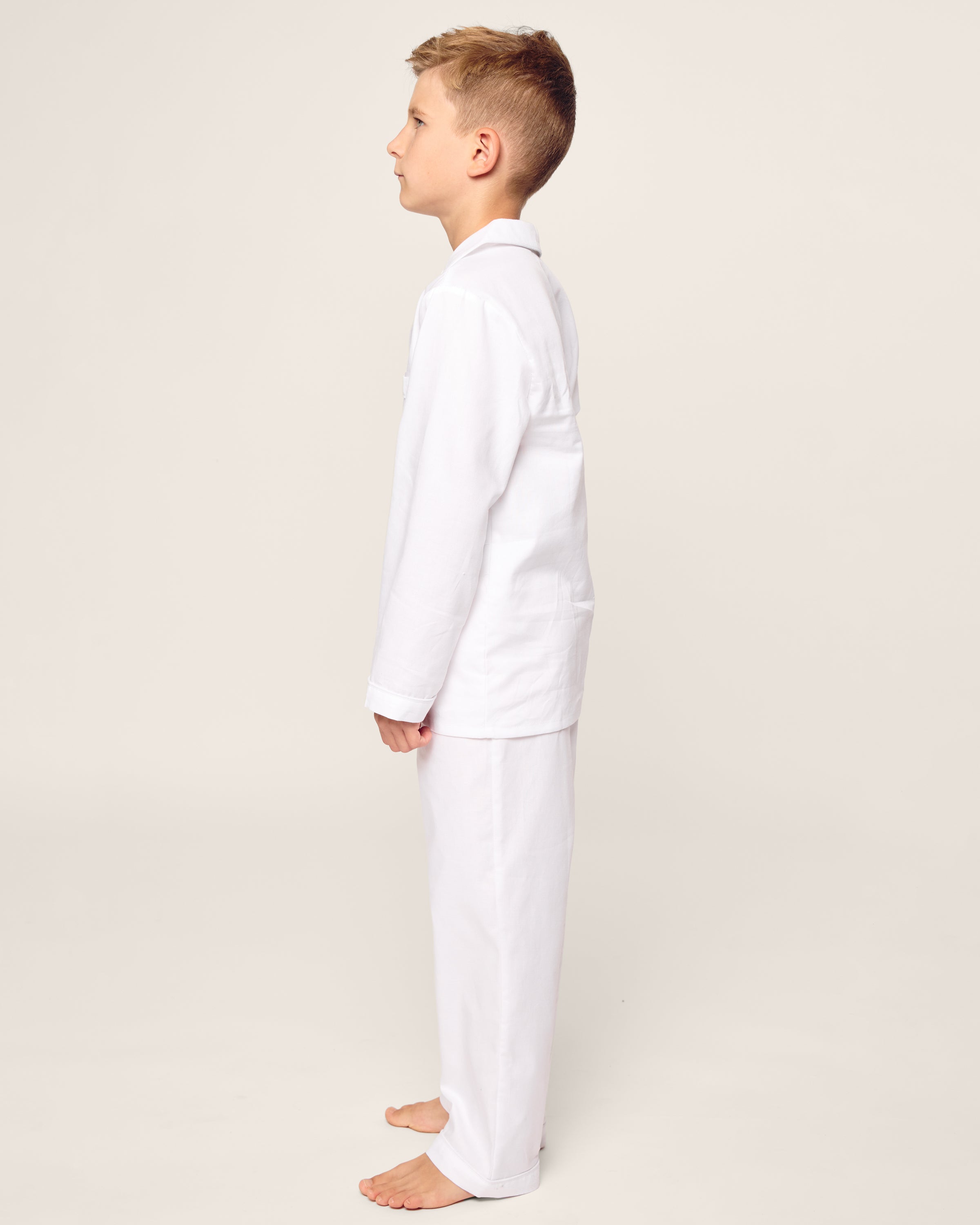 A young boy stands in profile, reminiscent of Petite Plumes Kids Twill Pajama Set. Barefoot against a plain, light background, he wears a white martial arts uniform, exuding focus as he gazes straight ahead.