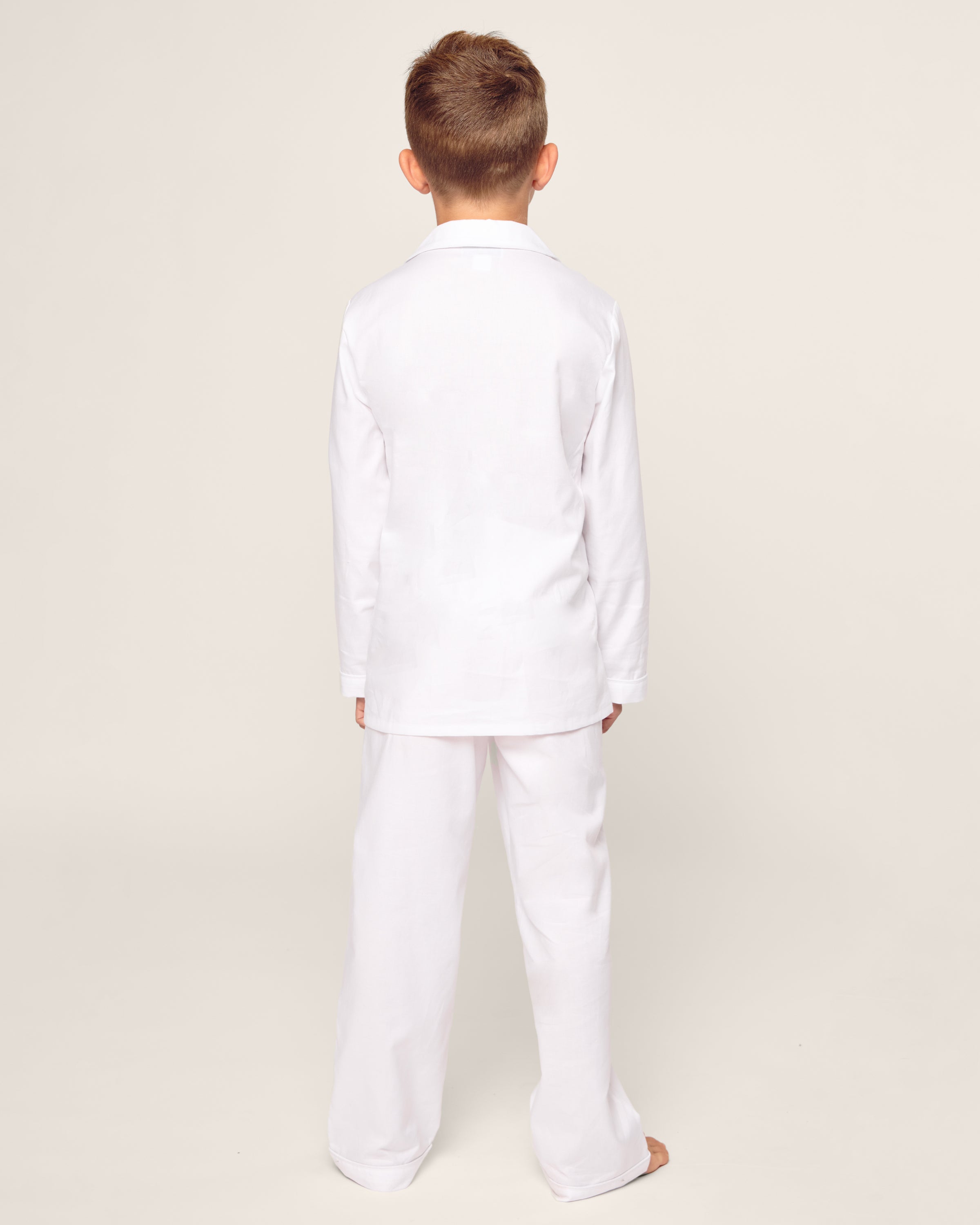 A child with short brown hair stands barefoot, facing away from the camera, wearing Petite Plumes luxurious Kids Twill Pajama Set in White with White Piping, complete with a long-sleeve shirt and matching pants, against a plain light background.