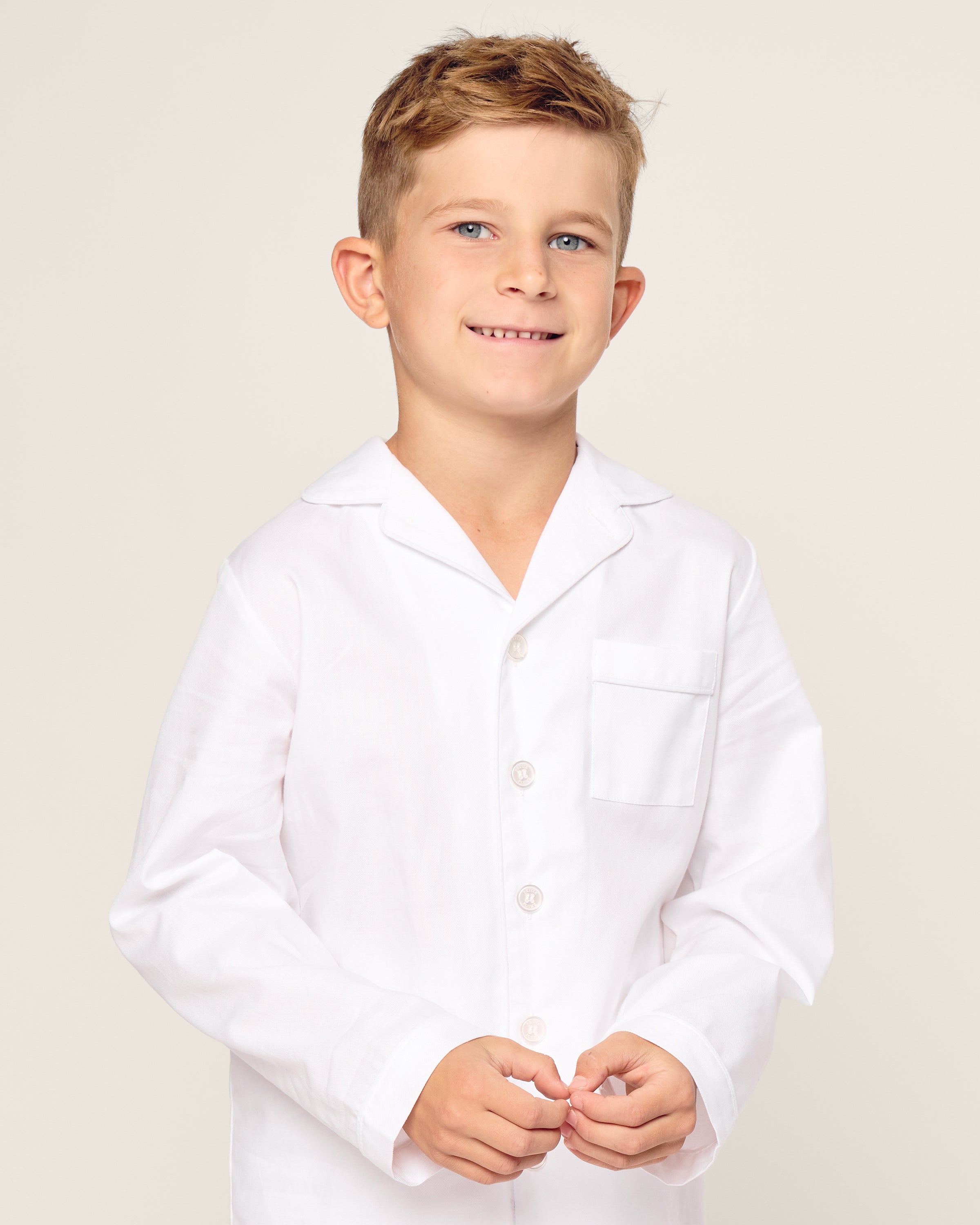 A young boy smiles warmly, wearing the Petite Plume Kids Twill Pajama Set in white with white piping. The crisp shirt features buttons and a chest pocket, ideal for luxurious sleepwear. He stands confidently against a plain, light-colored background.