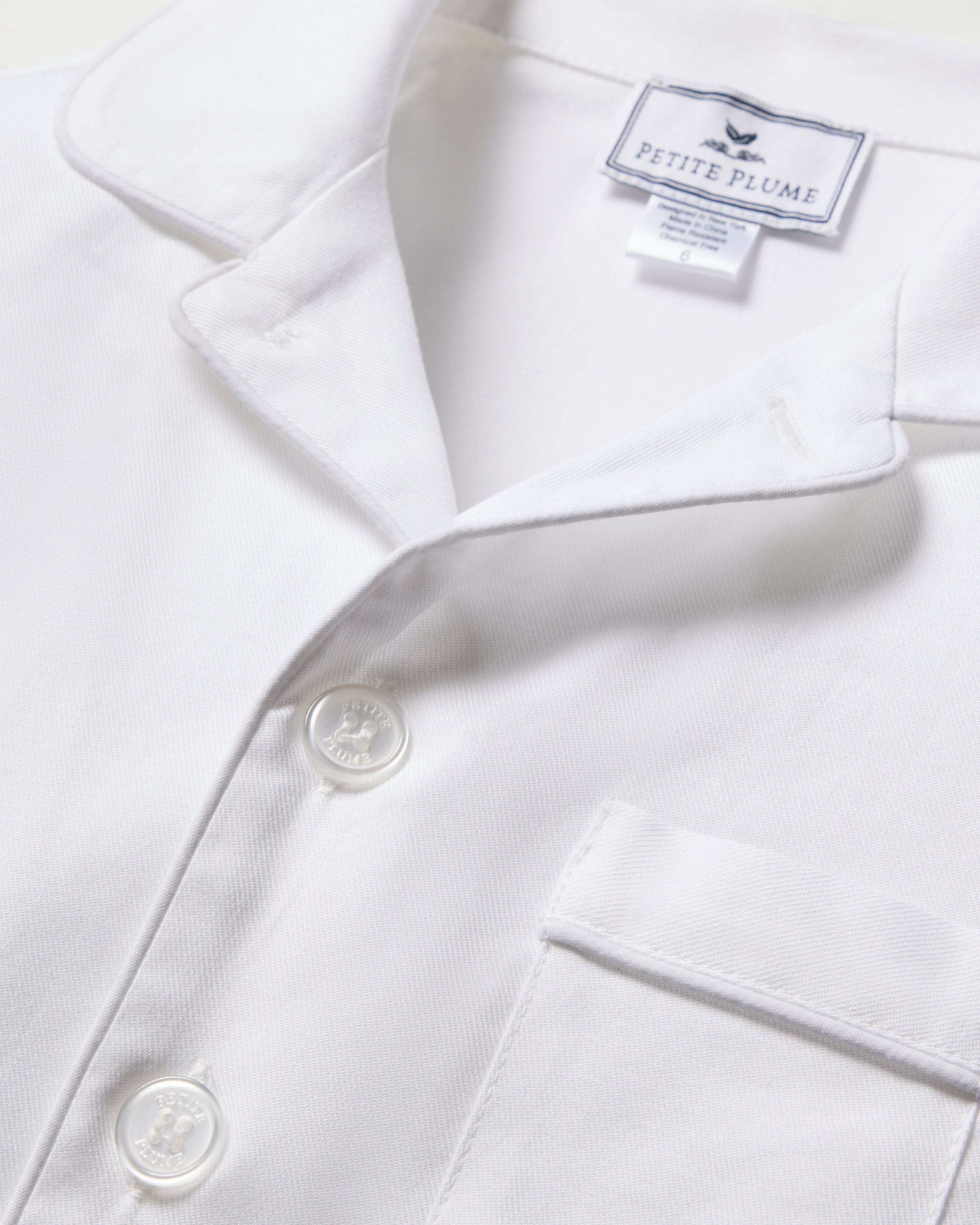 Close-up of the Kids Twill Pajama Set in White with Piping from Petite Plume, featuring three white buttons, a small left-side pocket, and a charming label illustration. The set offers textured fabric and a classic collar, ideal for restful nights.