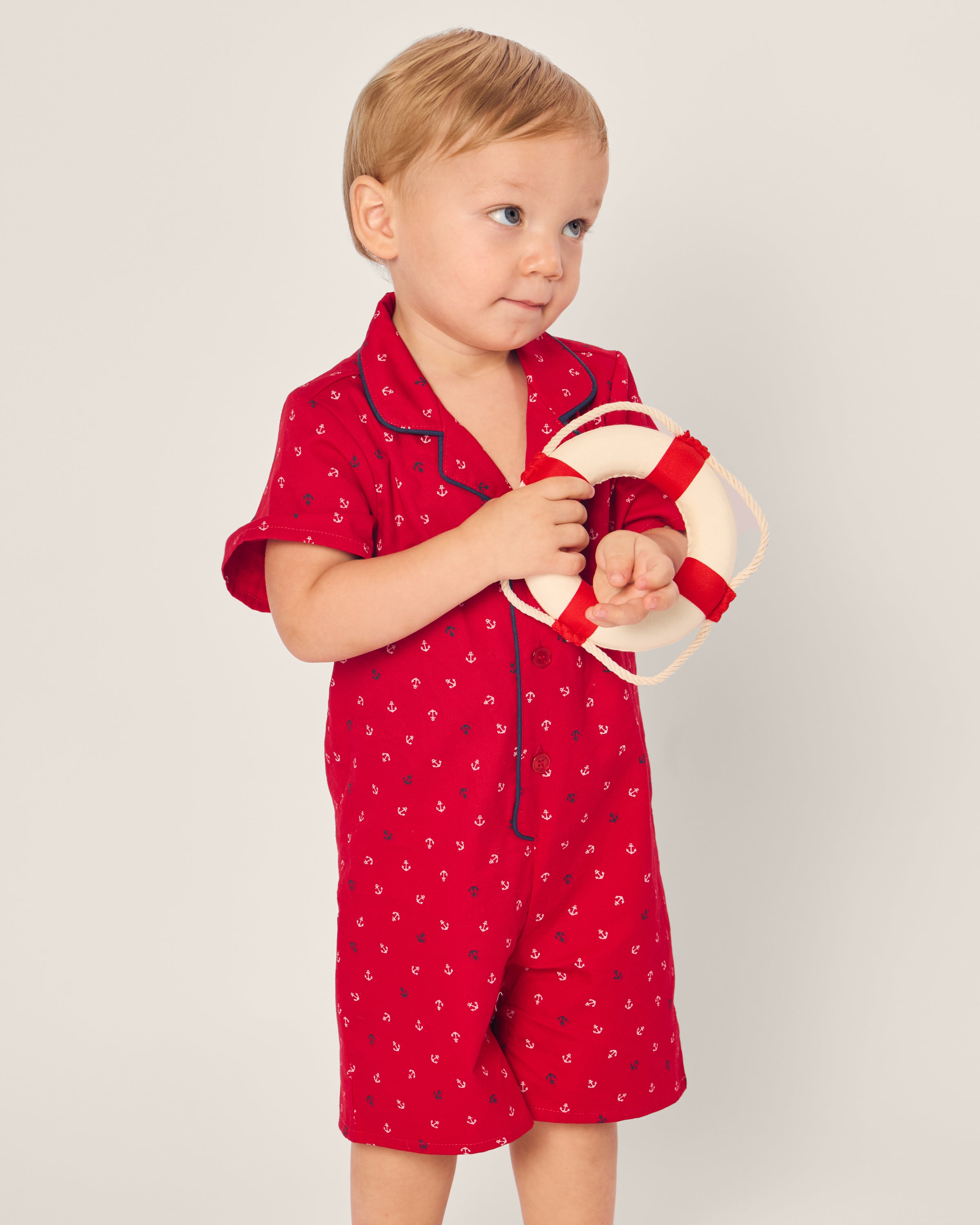 A toddler in the Petite Plume Babys Twill Summer Romper, featuring the Anchors Away print, holds a small life preserver. The child smiles subtly while looking to the side against a plain backdrop.