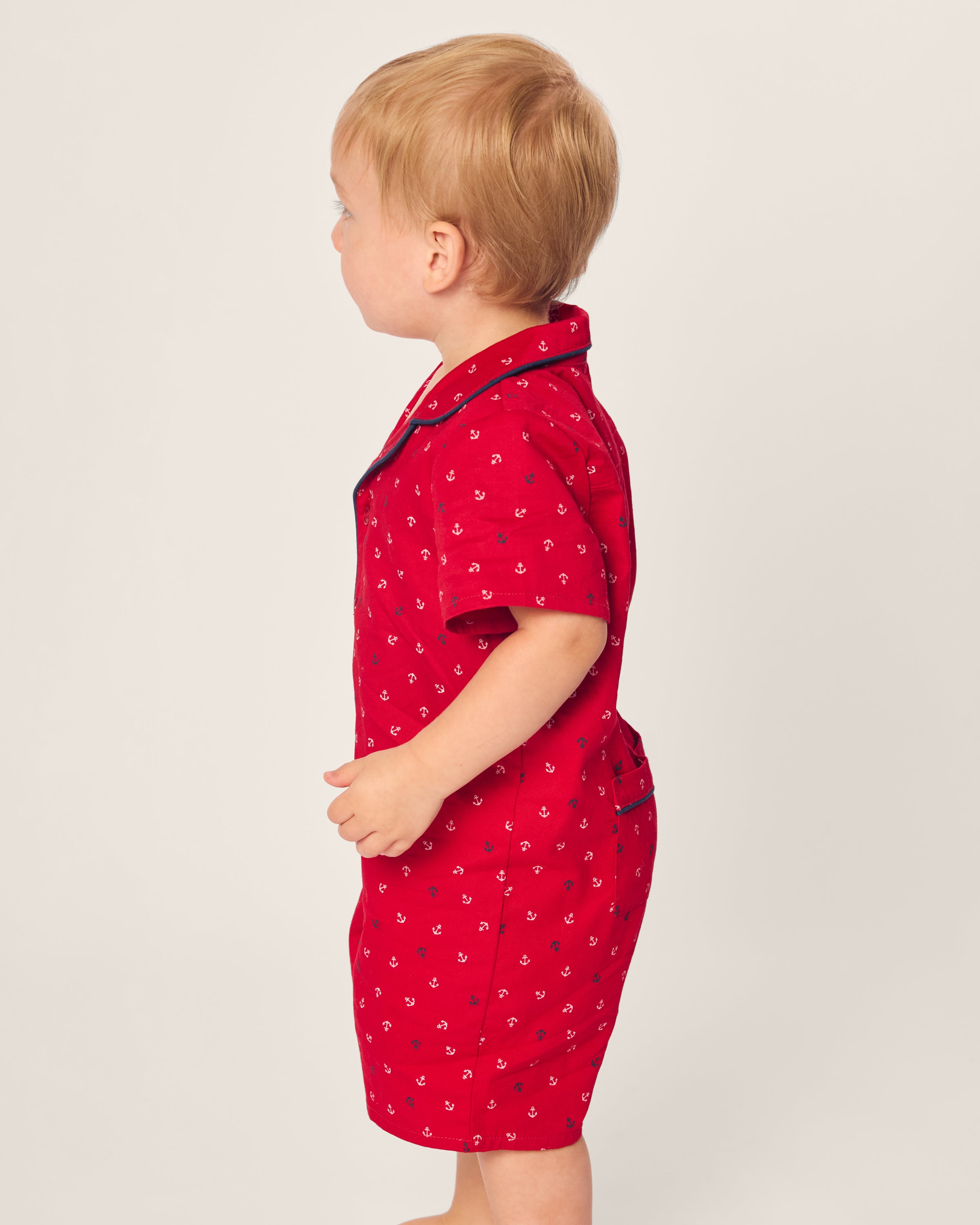 A toddler with light brown hair stands sideways in a Petite Plume Babys Twill Summer Romper featuring a red Anchors Away pattern. The flame-retardant sleepwear includes short sleeves, shorts, and dark trim at the collar and hem against a plain background.