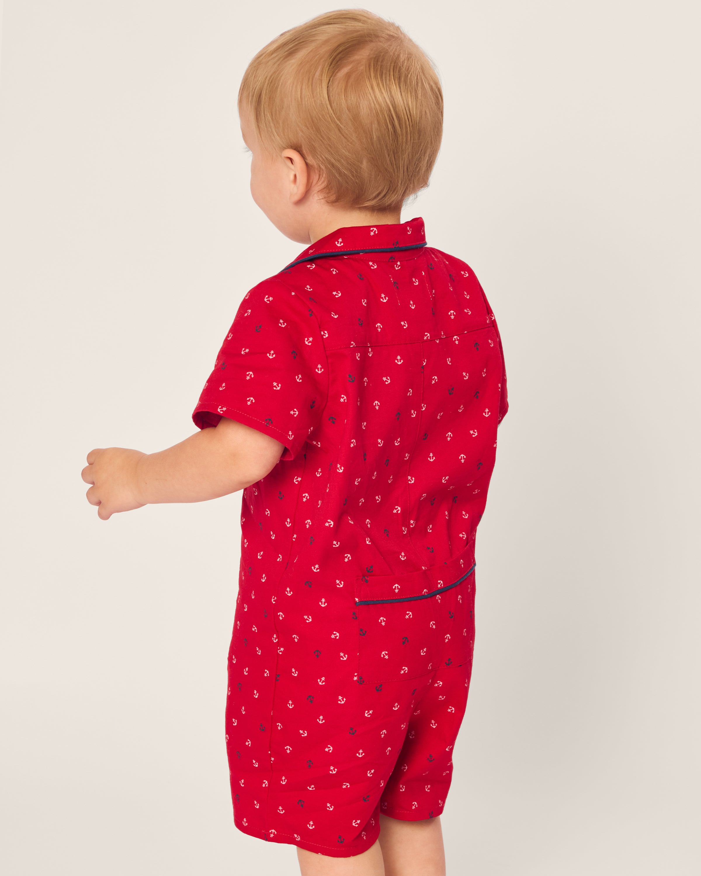 A toddler with light brown hair is seen from behind wearing Petite Plumes Babys Twill Summer Romper in Anchors Away. This red short-sleeve romper features small anchor patterns and a soft brushed twill fabric, set against a plain light background.