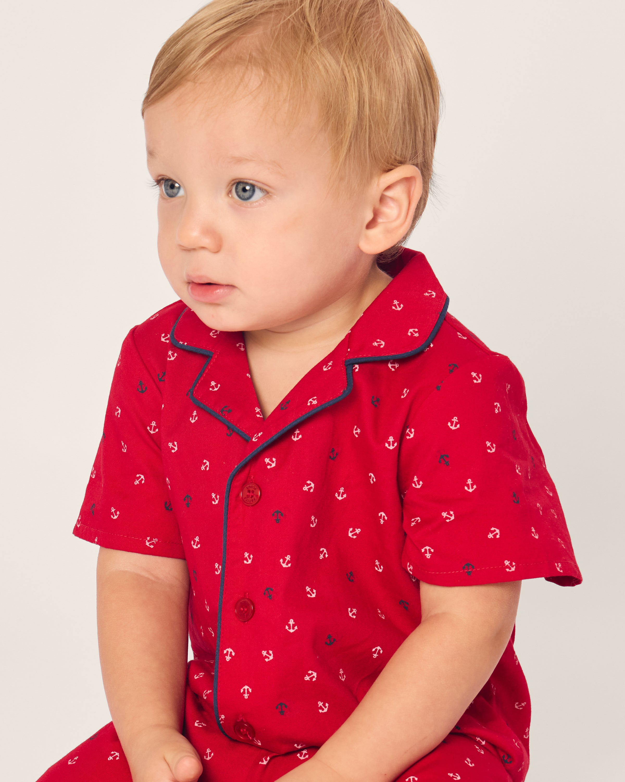 A toddler with light blond hair in Petite Plume’s Babys Twill Summer Romper in Anchors Away print sits against a neutral background. The flame retardant sleepwear has dark blue trim on the collar and button-down front.