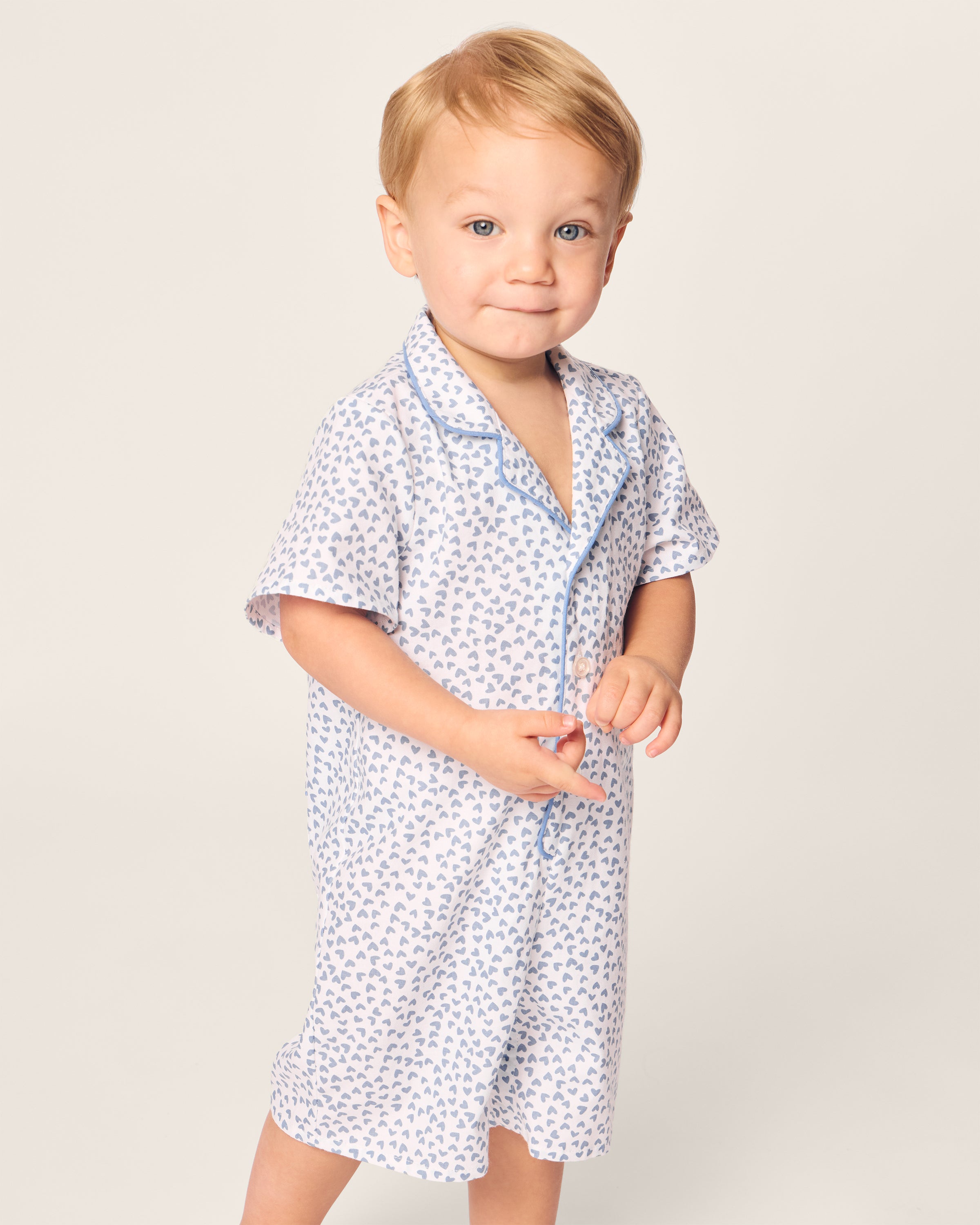 A young child stands against a plain background, smiling gently with short light hair and hands clasped, wearing Petite Plumes Babys Twill Romper featuring a blue and white Bluehearts design.