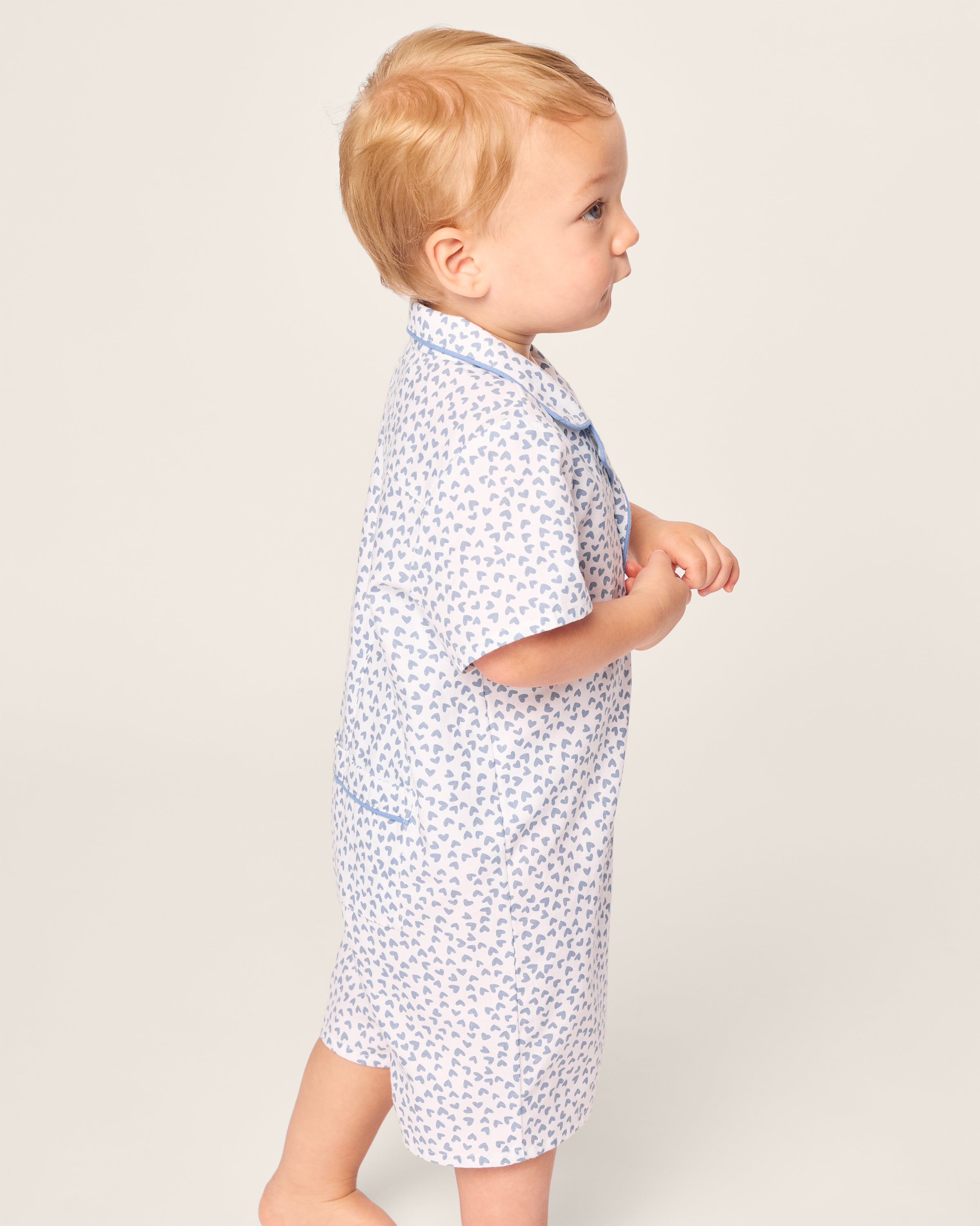 A toddler in a Petite Plume Babys Twill Romper in Bluehearts, with short sleeves, stands sideways against a plain background. The child has light hair and appears to be looking forward, hands resting gently together.