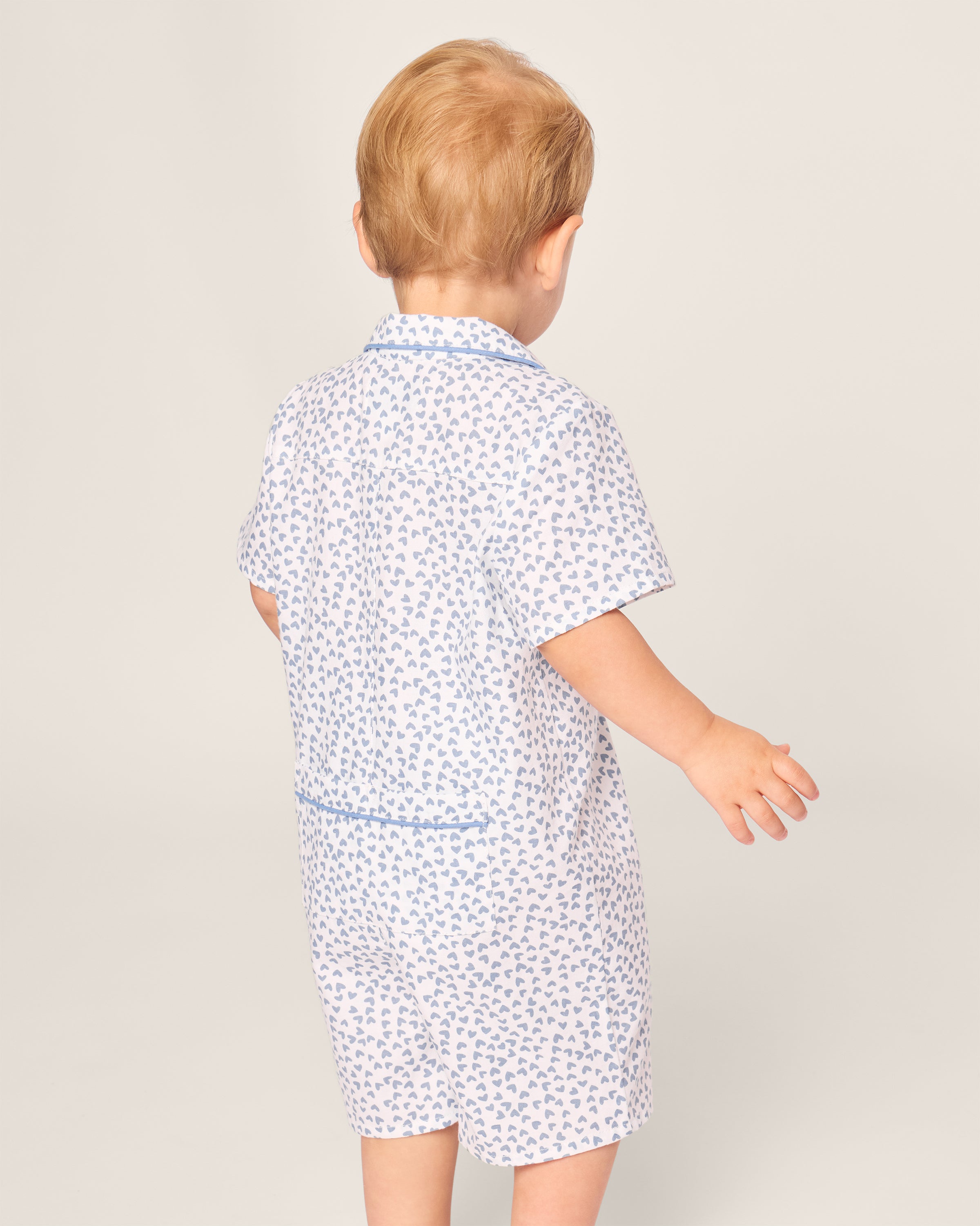 A light brown-haired toddler in Petite Plumes Bluehearts Babys Twill Romper stands against a plain background, facing away with outstretched arms.