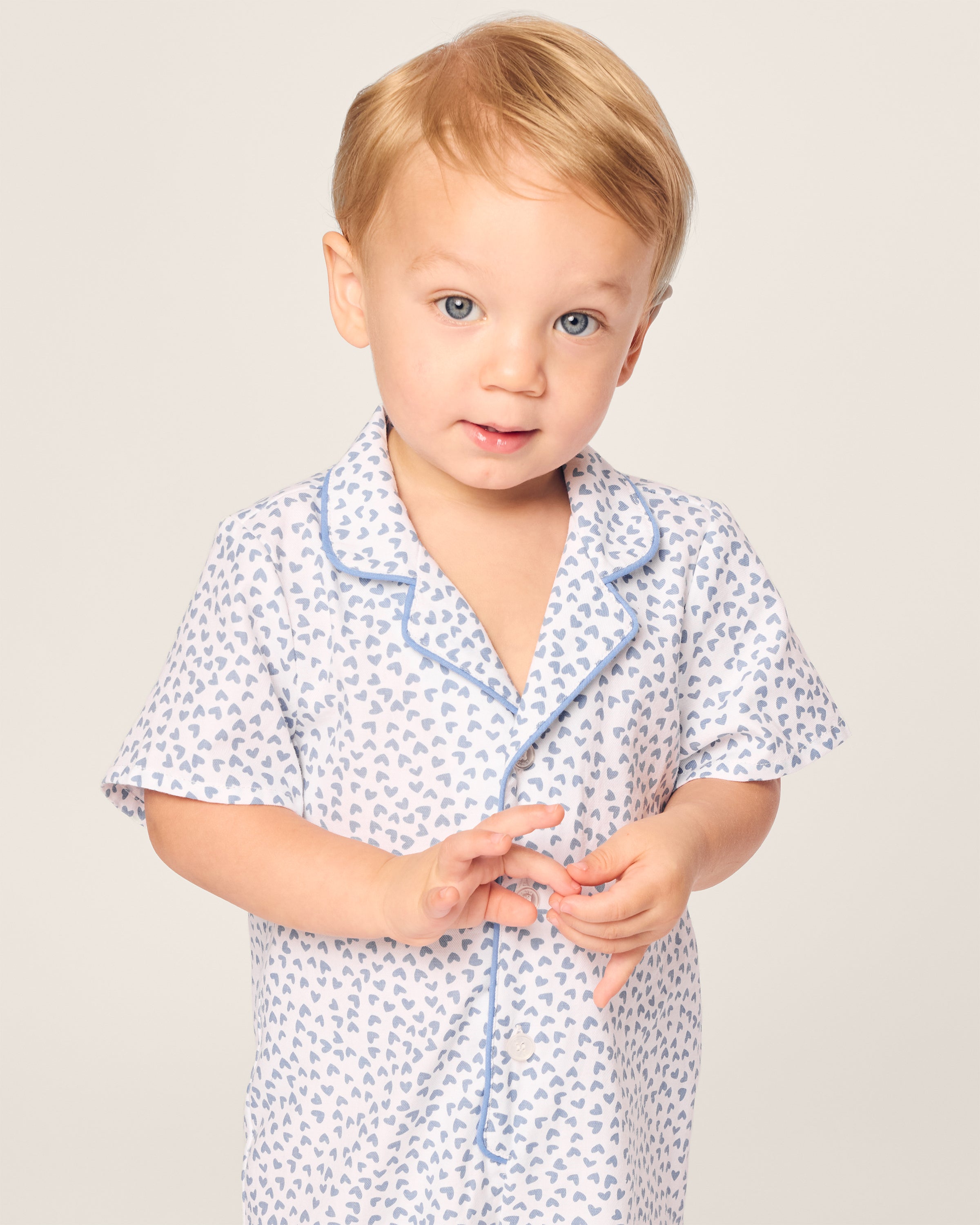 A toddler with blond hair wears Petite Plumes Babys Twill Romper in the charming Bluehearts design against a plain light background, their gentle expression perfectly complementing the subtle elegance of their attire.