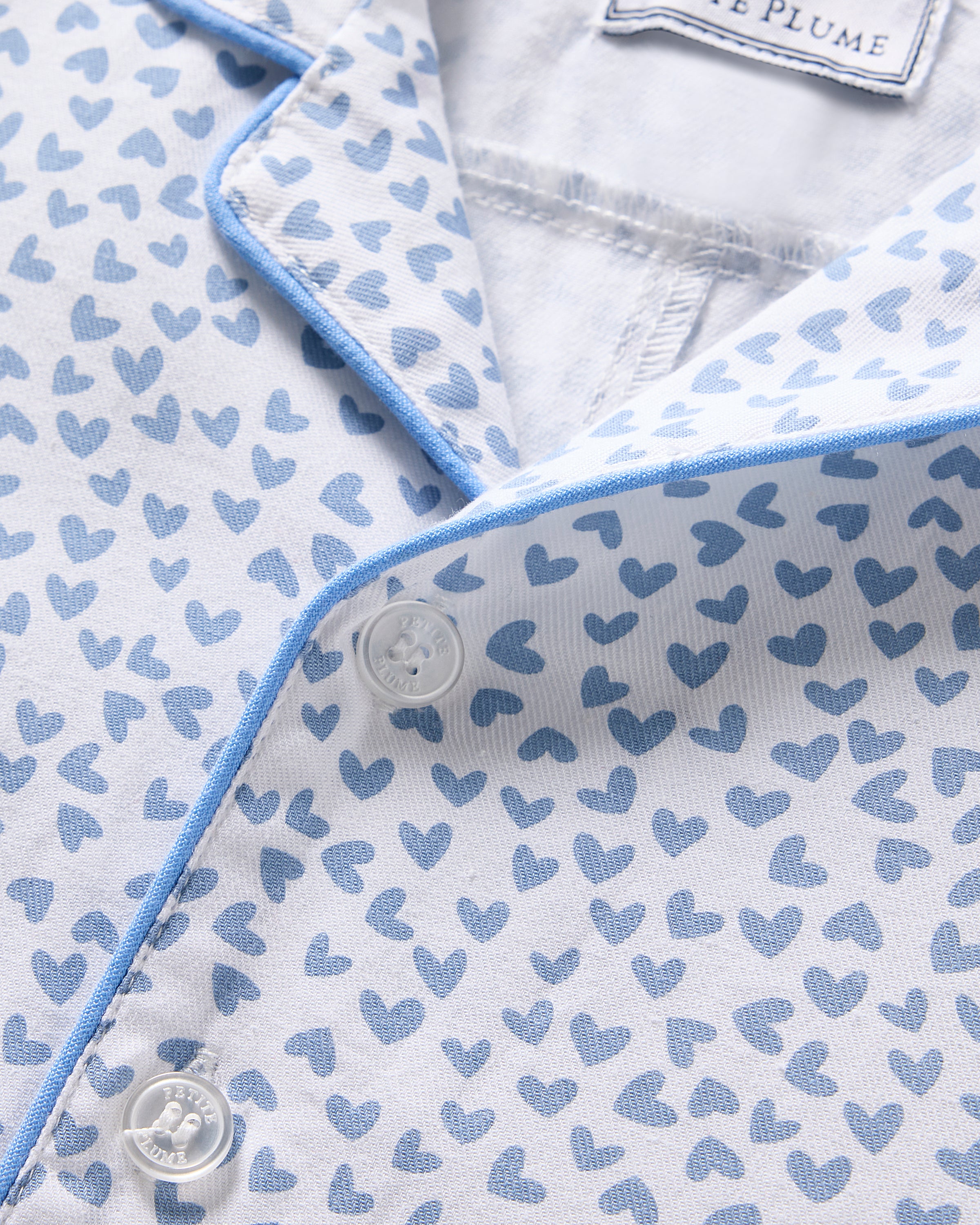 Close-up view of Petite Plumes Babys Twill Romper in Bluehearts, showcasing small blue hearts on white brushed twill. The design includes a blue-trimmed collar and placket with white buttons.