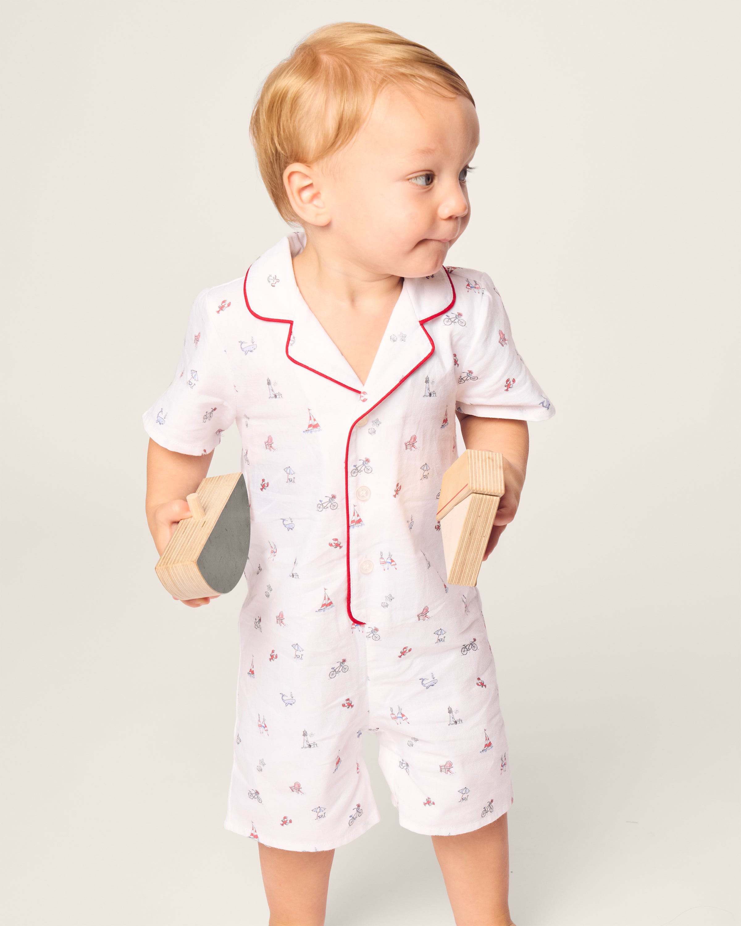 A toddler with light brown hair, dressed in Petite Plumes Babys Twill Summer Romper in Coastal Charms, featuring brushed twill fabric with red piping and playful prints, holds wooden blocks on a plain light background, radiating comfort and charm.