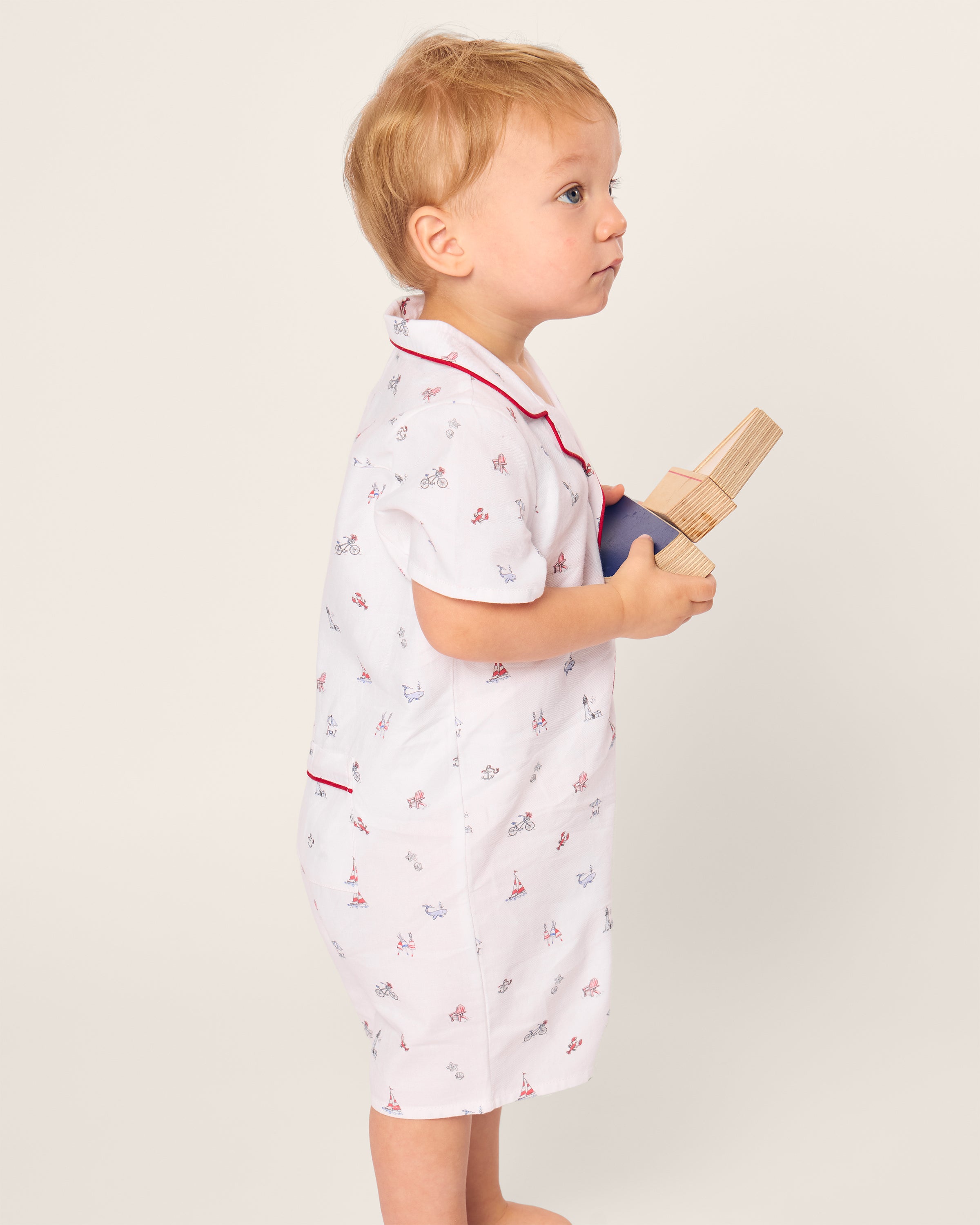 A toddler stands barefoot against a plain backdrop, donning Petite Plumes Babys Twill Summer Romper in Coastal Charms, featuring small designs and red trim. The brushed twill fabric complements the wooden blocks they hold while gazing attentively to the side.