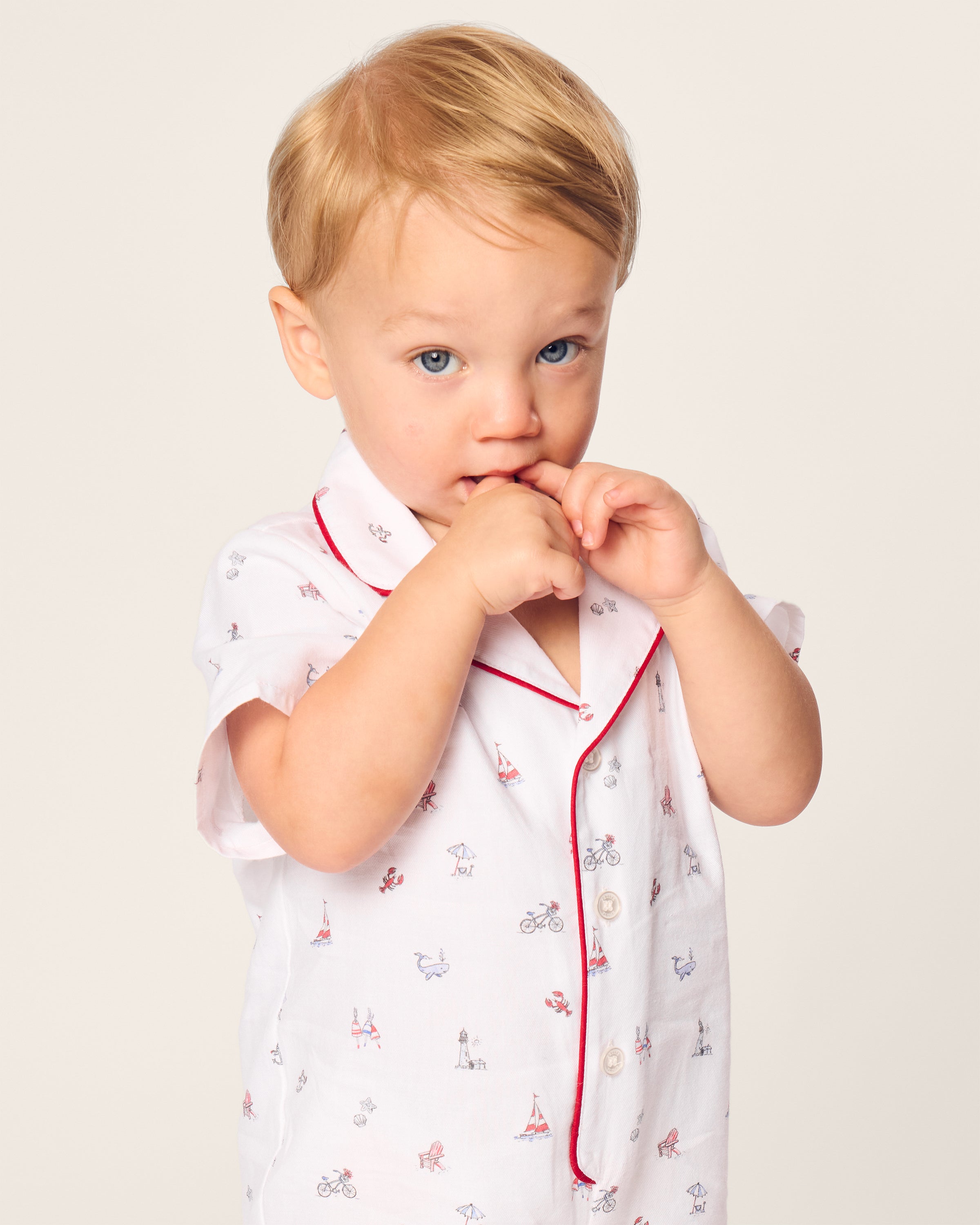 A blonde toddler wears a Petite Plume white Babys Twill Summer Romper in Coastal Charms, featuring playful animal and bicycle patterns and red piping. Made from cozy brushed twill, the child gazes thoughtfully at the camera with fingers touching their lips against a plain backdrop.