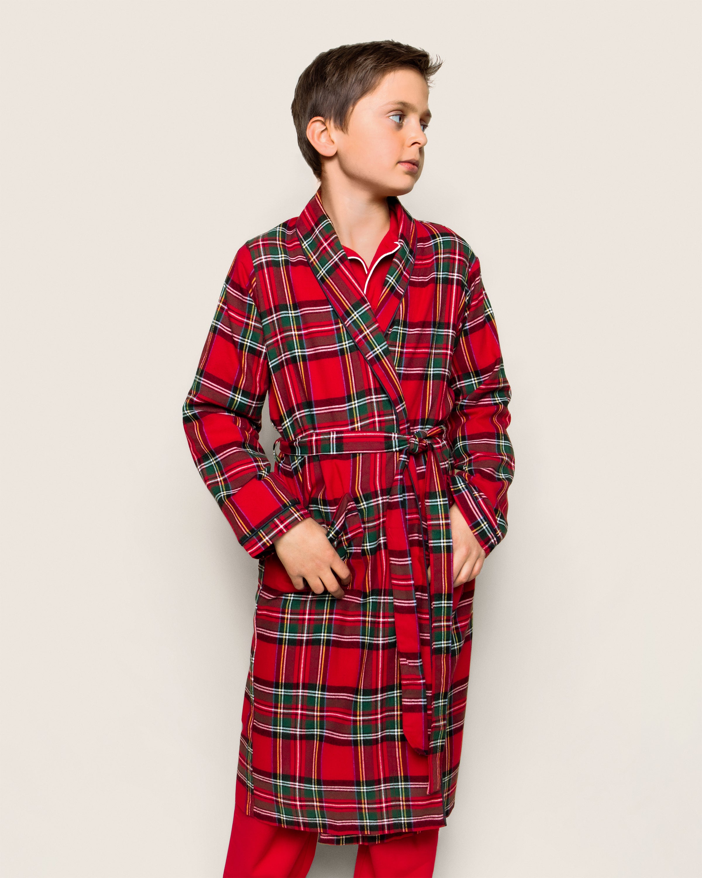 A young boy models Petite Plume’s Kids Brushed Cotton Robe in Imperial Tartan, featuring a belt and pockets. Made from lightweight soft flannel with a neutral expression, he stands before a plain, light background.