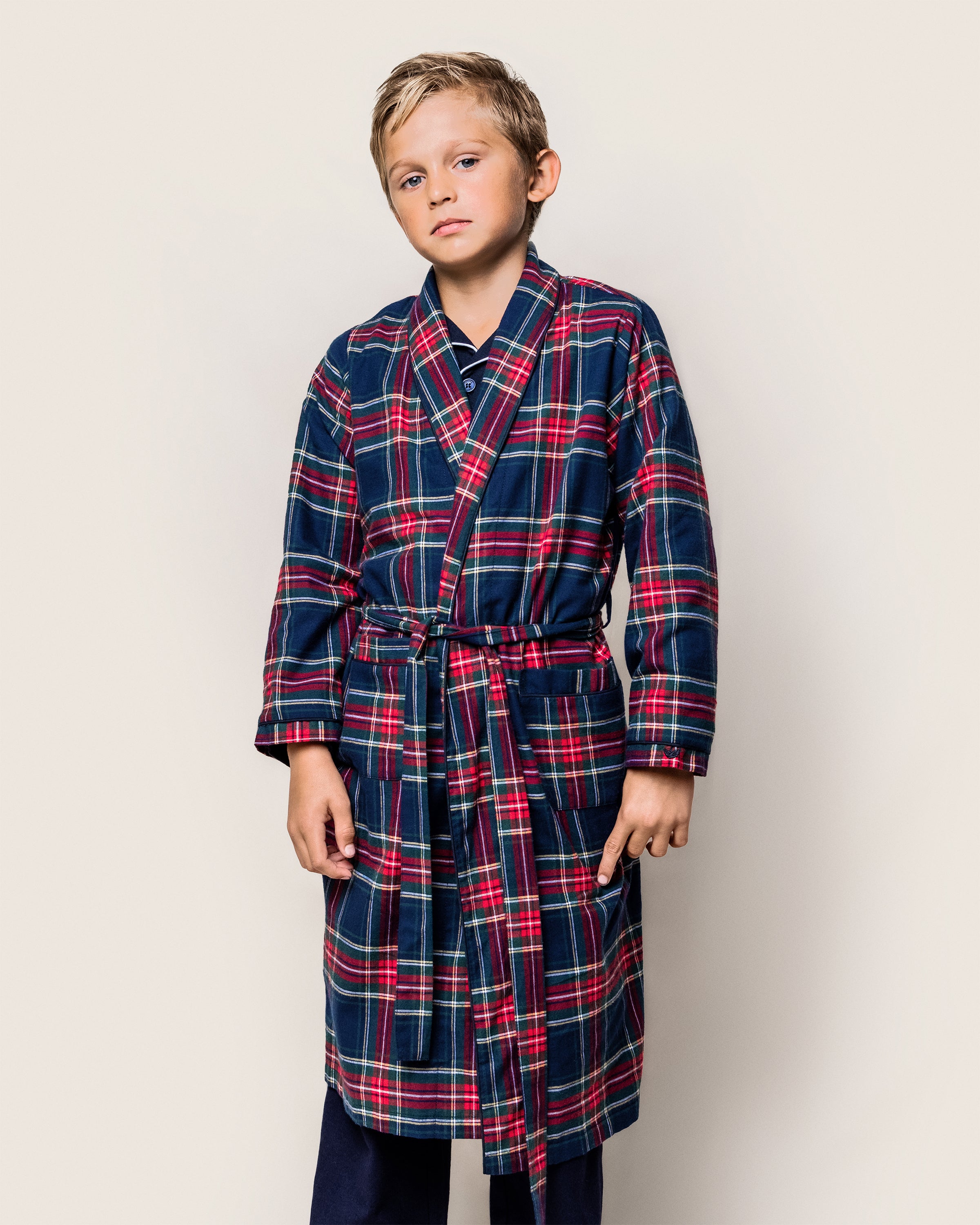 A young boy in a Petite Plume Kids Brushed Cotton Robe in Windsor Tartan stands against a plain background. With short blond hair and a neutral expression, his hands hang loosely by his sides, capturing the essence of cozy sleepwear.