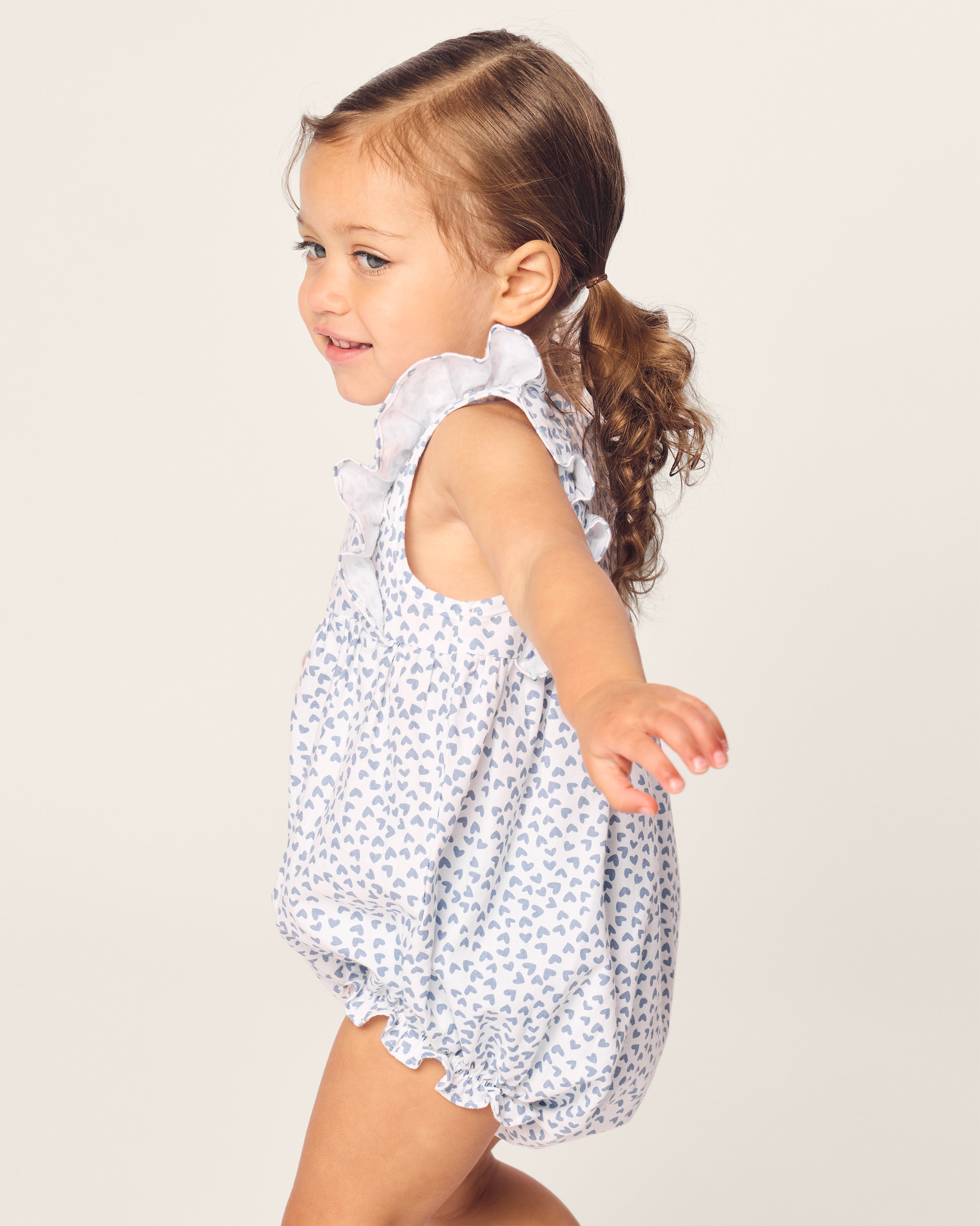 A brown-haired toddler with a ponytail smiles while wearing Petite Plumes Babys Twill Ruffled Romper in Bluehearts. The light-colored yarn-dyed cotton fabric and ruffled sleeves enhance her cheerful look against the plain, light background.