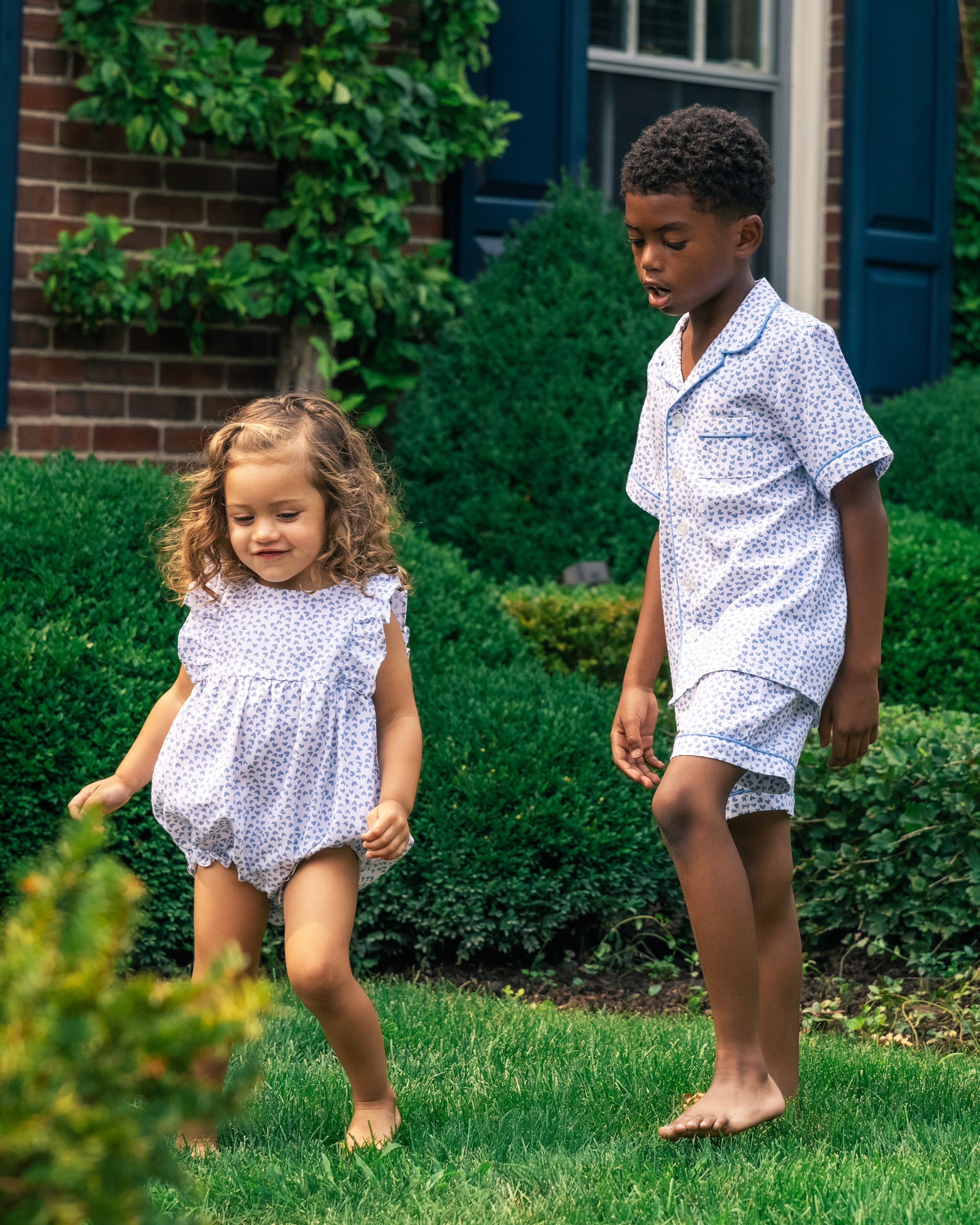 Two young children, a girl and a boy, play barefoot on a grassy lawn. They joyfully romp in coordinating Petite Plume outfits featuring Bluehearts designs with blue and white polka dots. Green shrubs and a brick wall with a window frame the background.