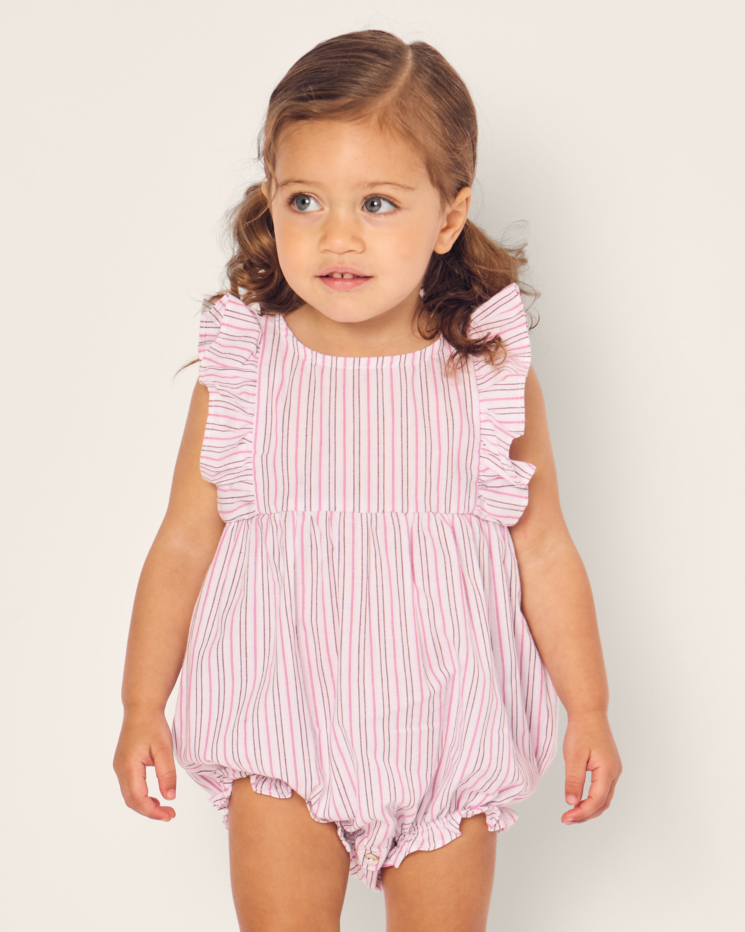 A toddler stands against a plain background, wearing Petite Plumes Babys Twill Ruffled Romper in Blush Boulevard. With ruffled sleeves and light brown hair in pigtails, the child gazes slightly to the side, capturing an adorable moment.