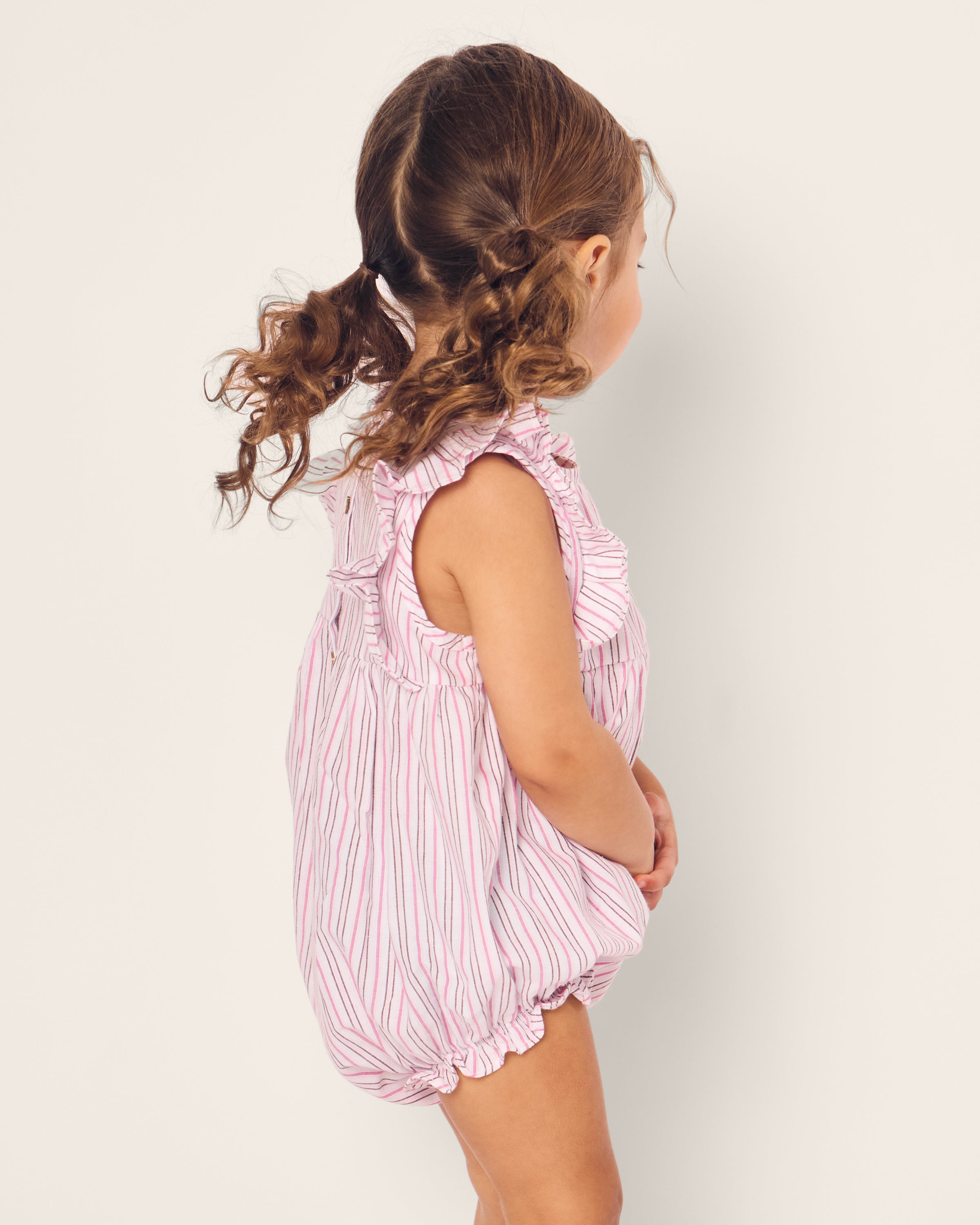 A young child with curly hair showcases Petite Plumes Babys Twill Ruffled Romper in Blush Boulevard. Facing sideways with hands clasped, they gaze downward slightly. The plain, light background emphasizes the charm of this cozy sleepwear ensemble.