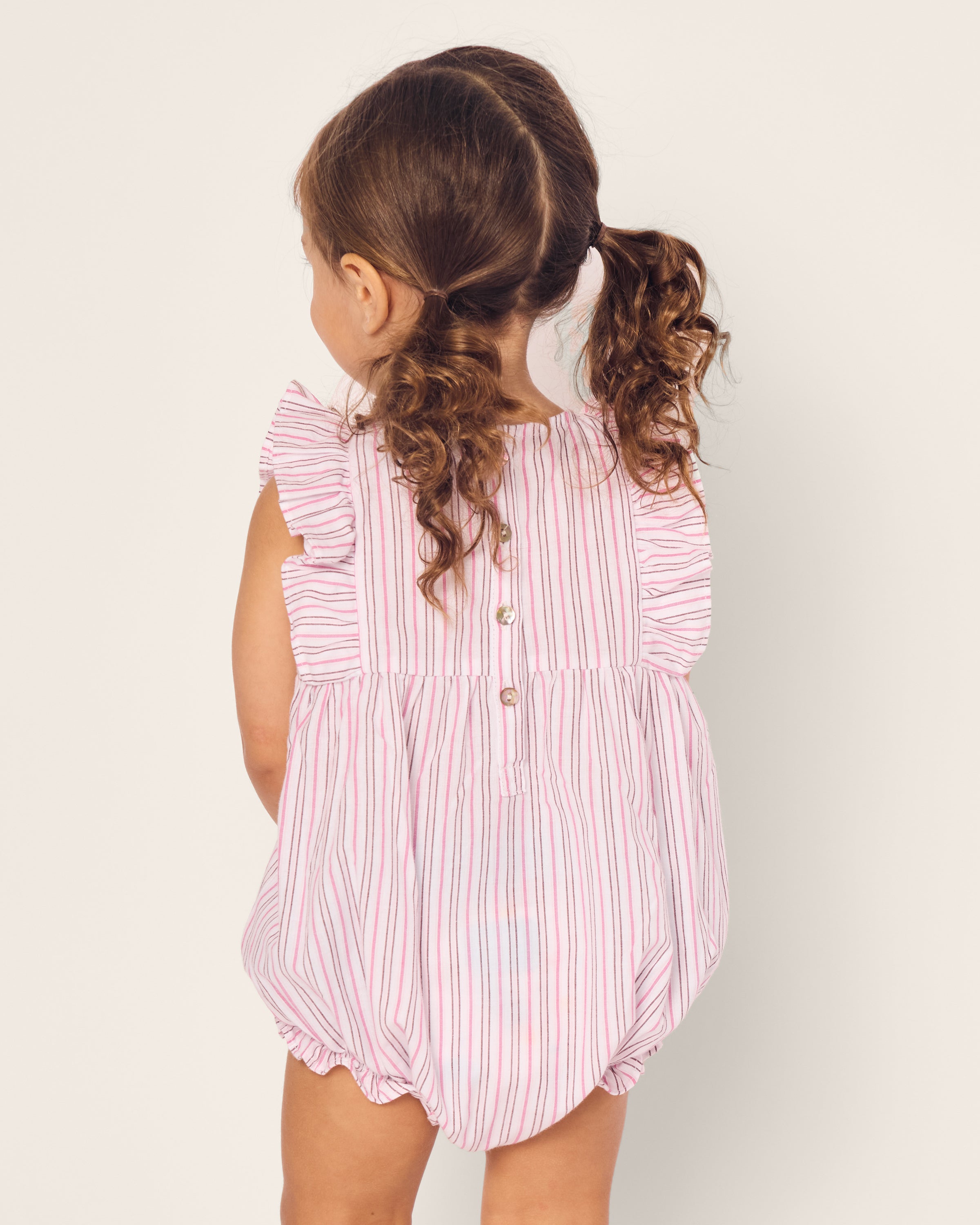 A toddler with curly brown hair in pigtails is wearing Petite Plumes Babys Twill Ruffled Romper in Blush Boulevard. Crafted for comfort, the romper has buttons on the back and ruffled sleeves. The child is facing away from the camera against a simple light background.