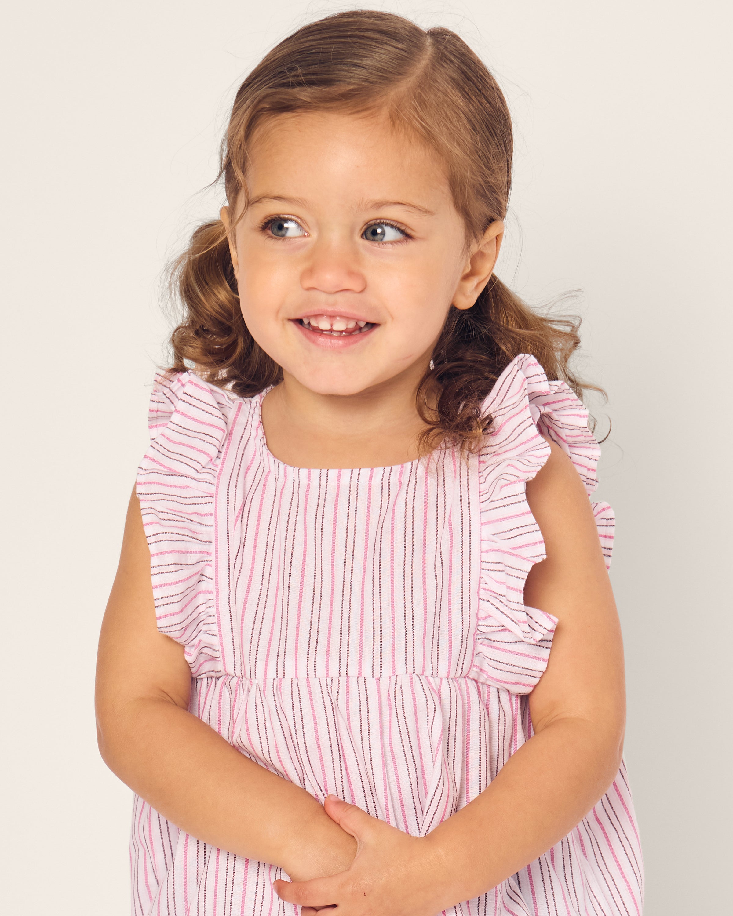 A brown-haired toddler with pigtails smiles while wearing Petite Plumes Babys Twill Ruffled Romper in pink and white stripes from Blush Boulevard, set against a plain background.