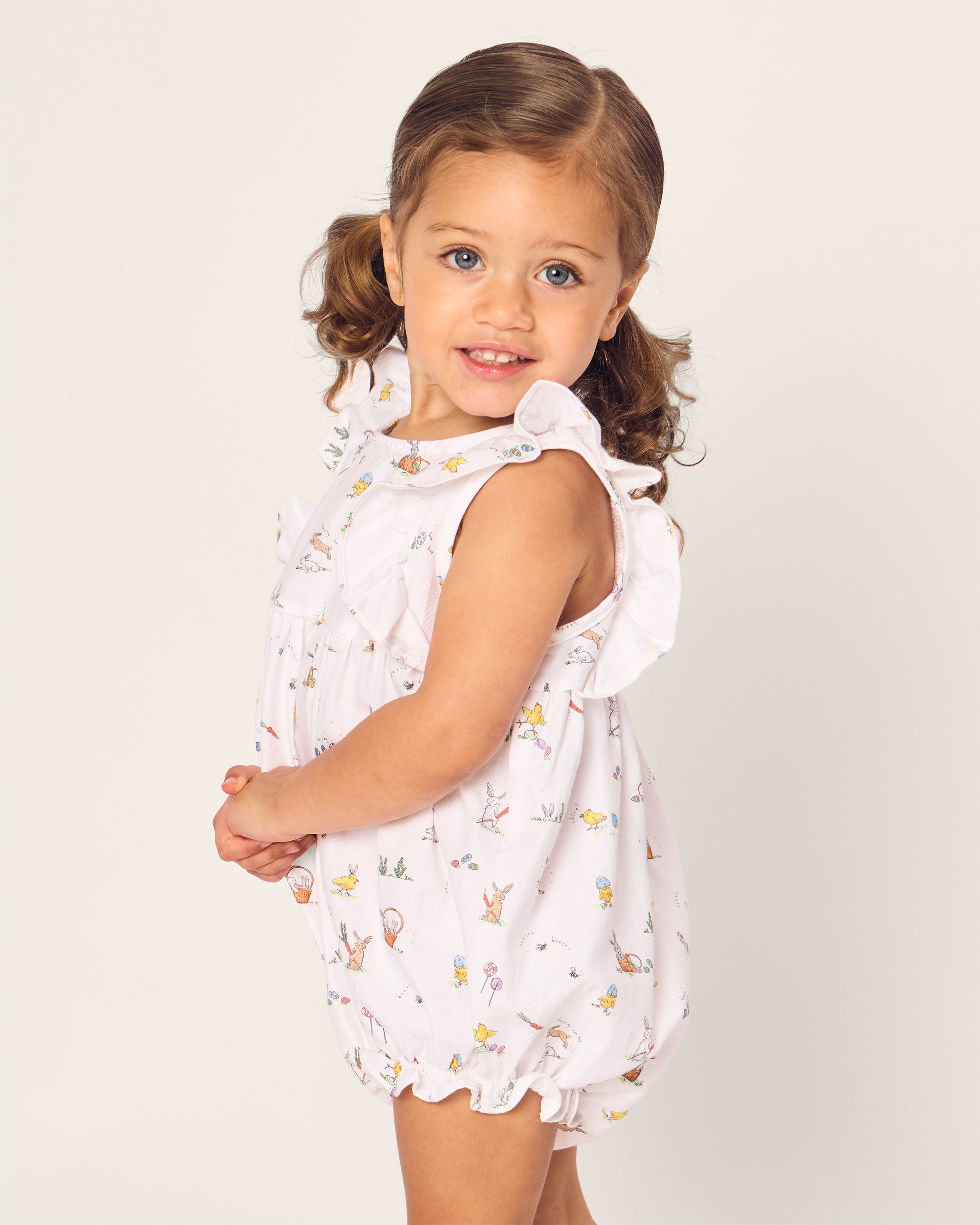 A toddler with brown pigtails smiles at the camera, wearing Petite Plumes Babys Twill Ruffled Romper in Easter Frolic. The romper features ruffled sleeves, and a plain, light-colored background highlights her playful innocence.