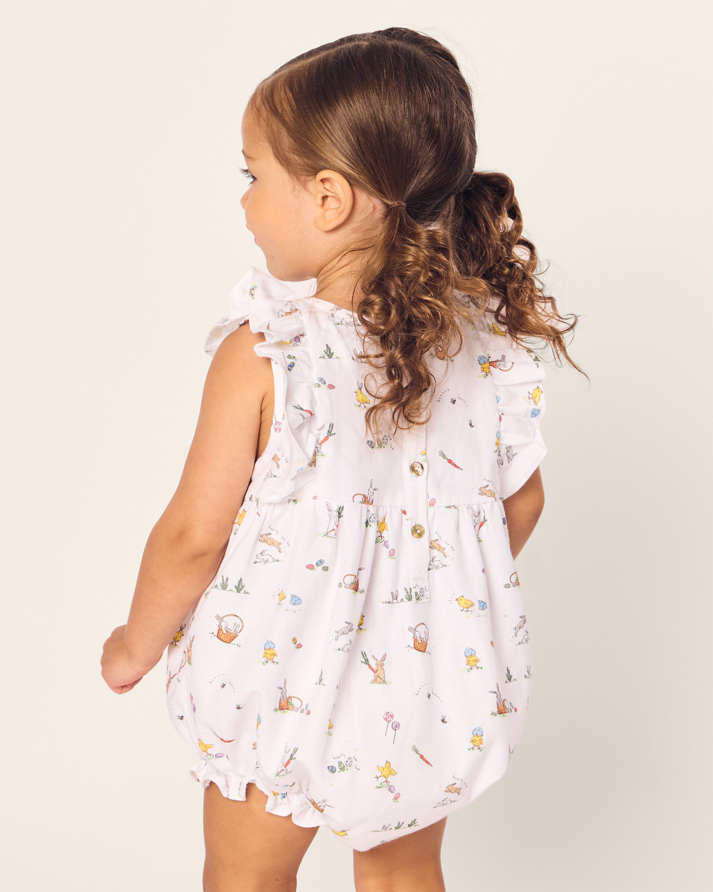 A young child with curly hair in a ponytail wears Petite Plumes Babys Twill Ruffled Romper in Easter Frolic. The sleeveless, white romper features ruffled edges and stands out against a plain, light background as the child faces away from the camera.