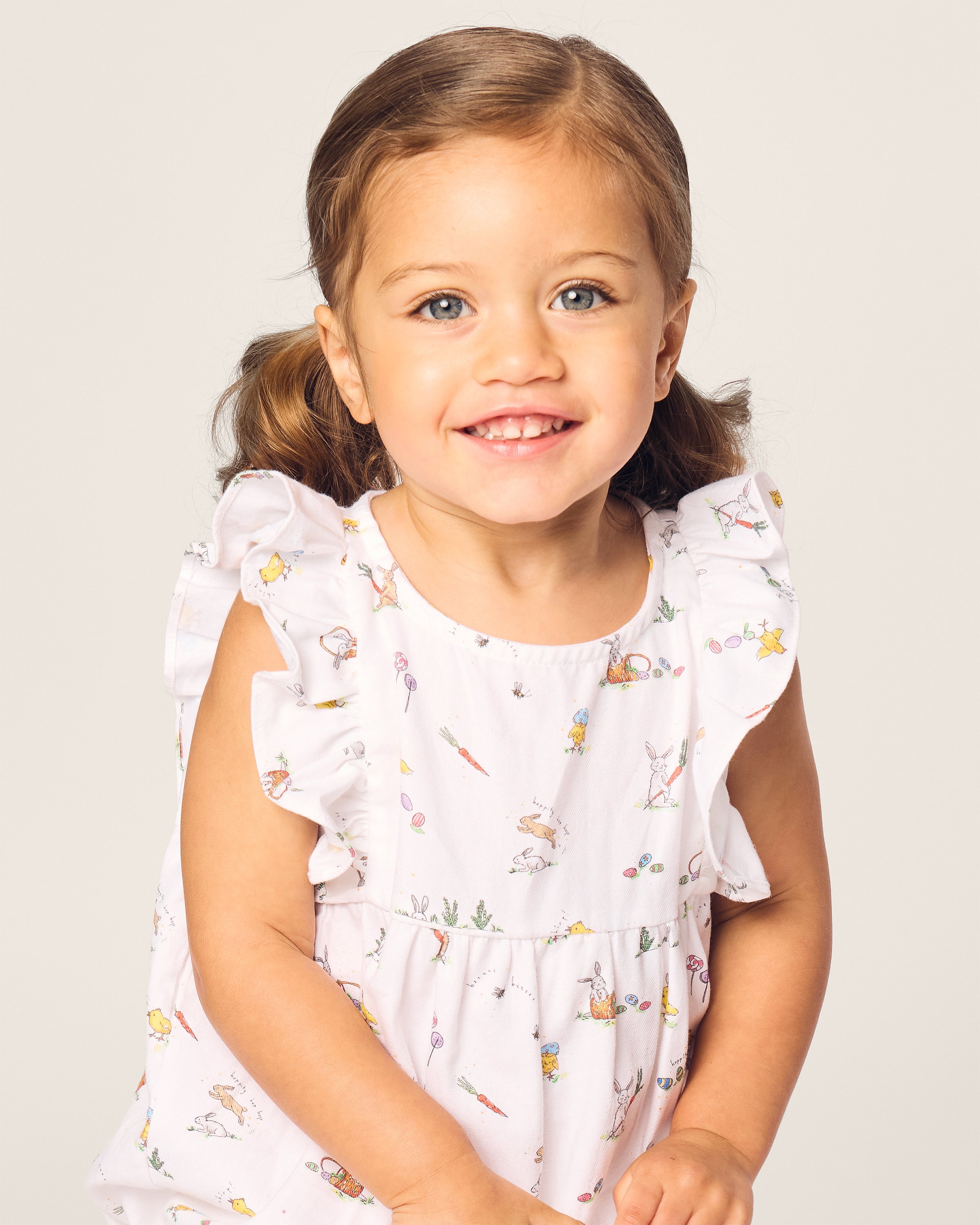 A young child with brown hair in pigtails, wearing Petite Plumes Babys Twill Ruffled Romper in Easter Frolic, smiles at the camera against a plain background.