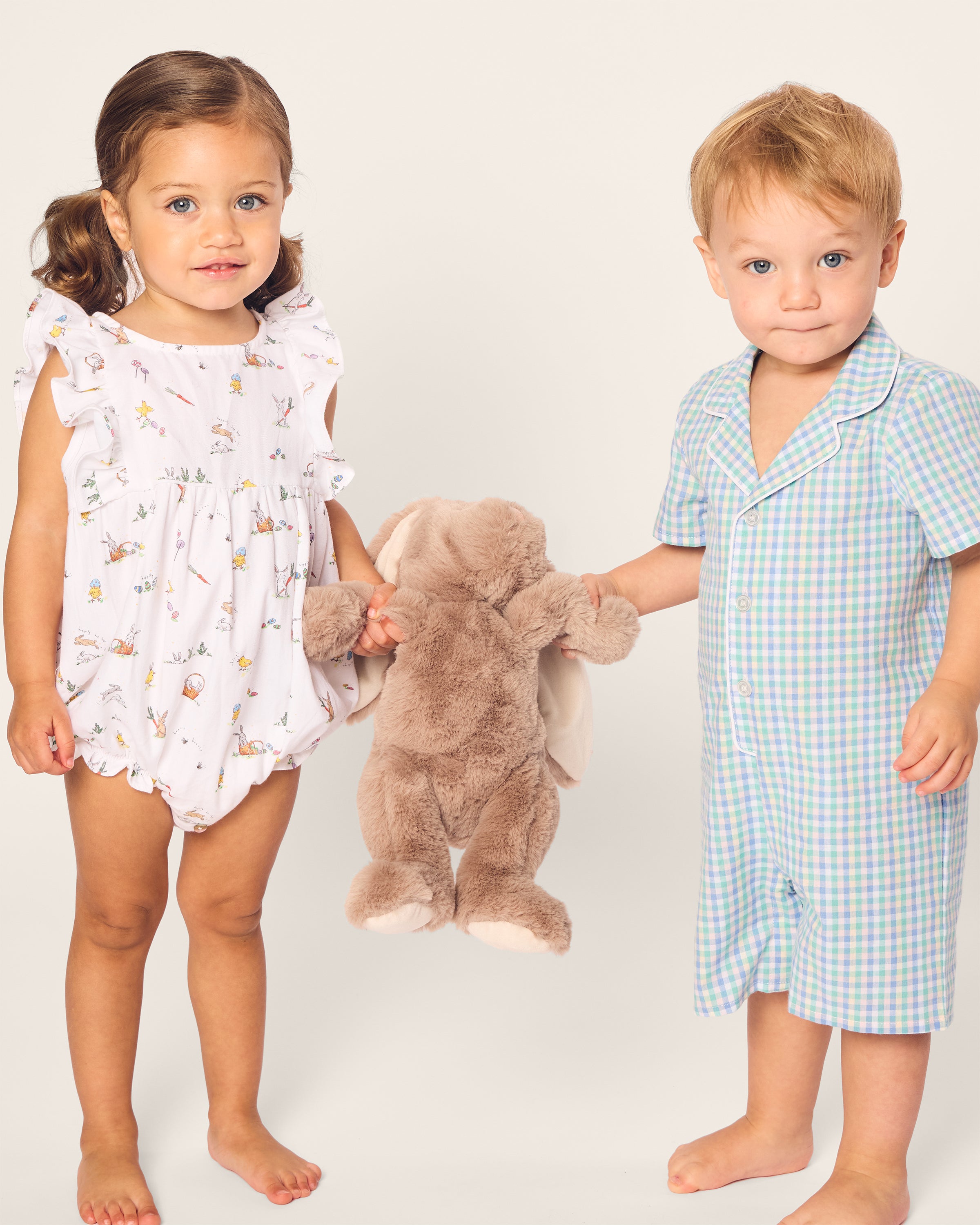 A young girl with pigtails in a Petite Plume Babys Twill Ruffled Romper in Easter Frolic and a boy with short blond hair in matching sleepwear stand together holding a plush bunny against a plain background.