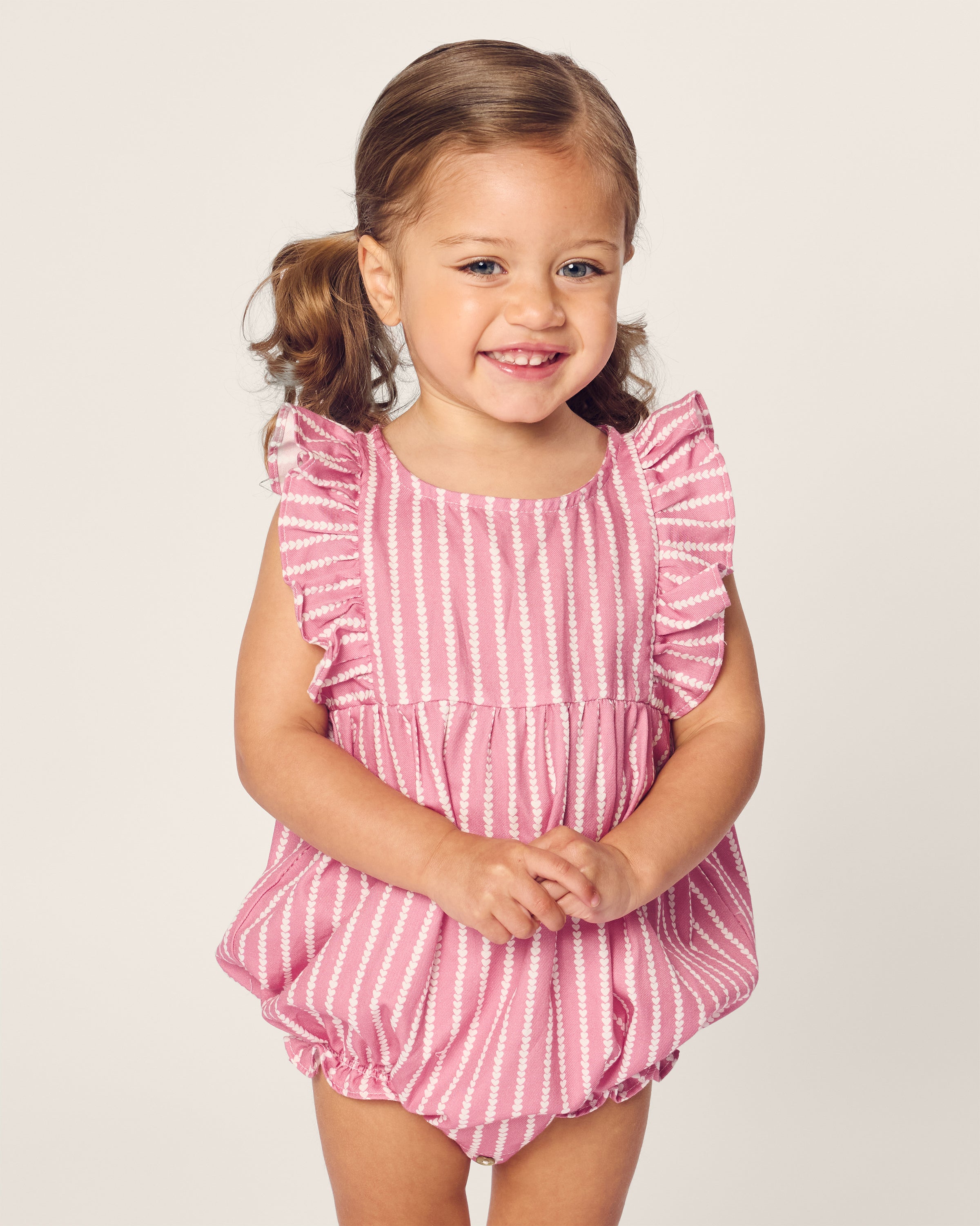 A young child beams in Petite Plumes Babys Twill Ruffled Romper in Love Lines, her brown hair tied in pigtails. The simple yet charming style evokes a Victorian era feel, standing out against the light background.