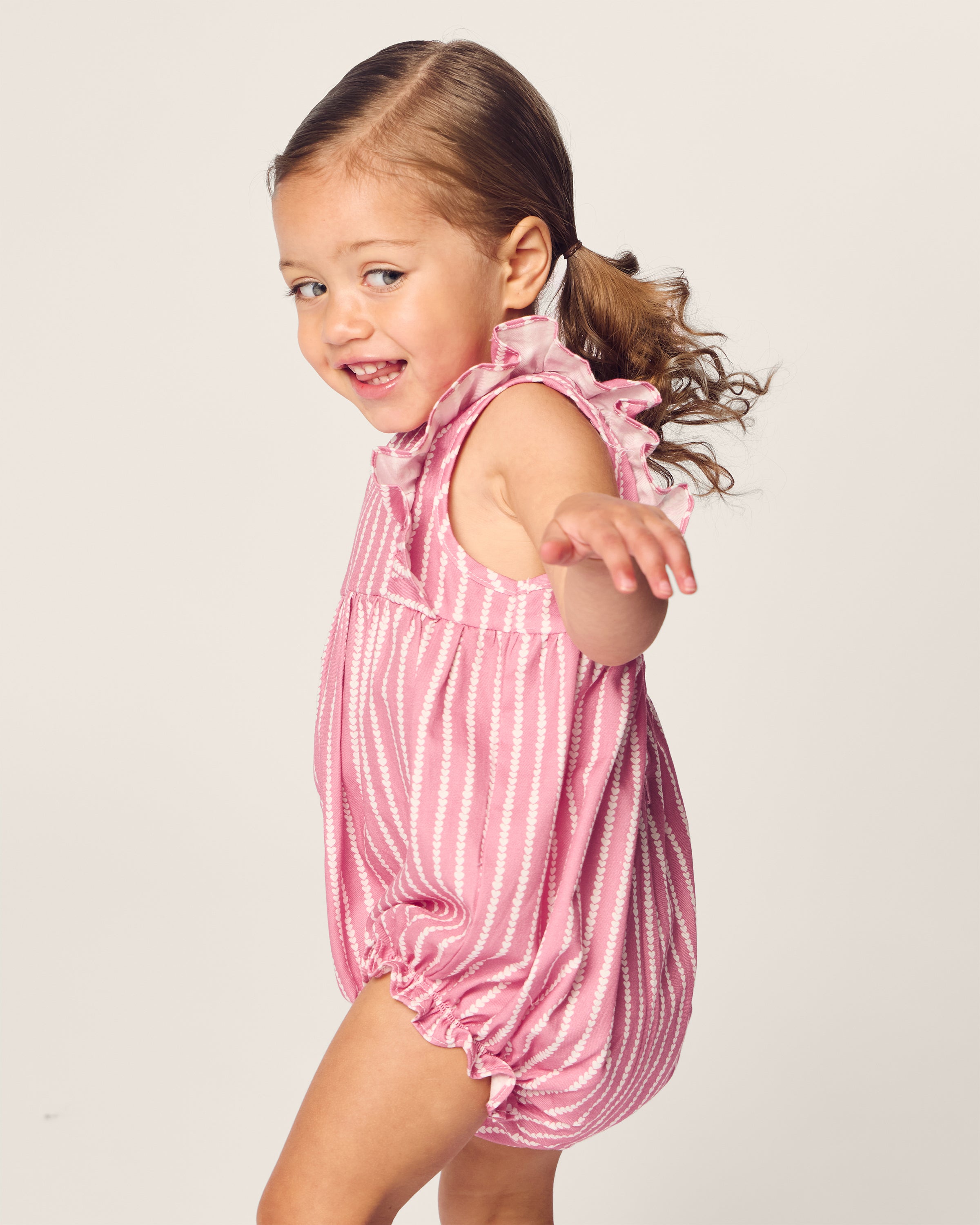 A young child with brown hair in a ponytail wears Petite Plumes delightful Babys Twill Ruffled Romper in Love Lines, perfect for cozy evenings. Smiling and facing sideways with one arm extended, the background is a plain light color.