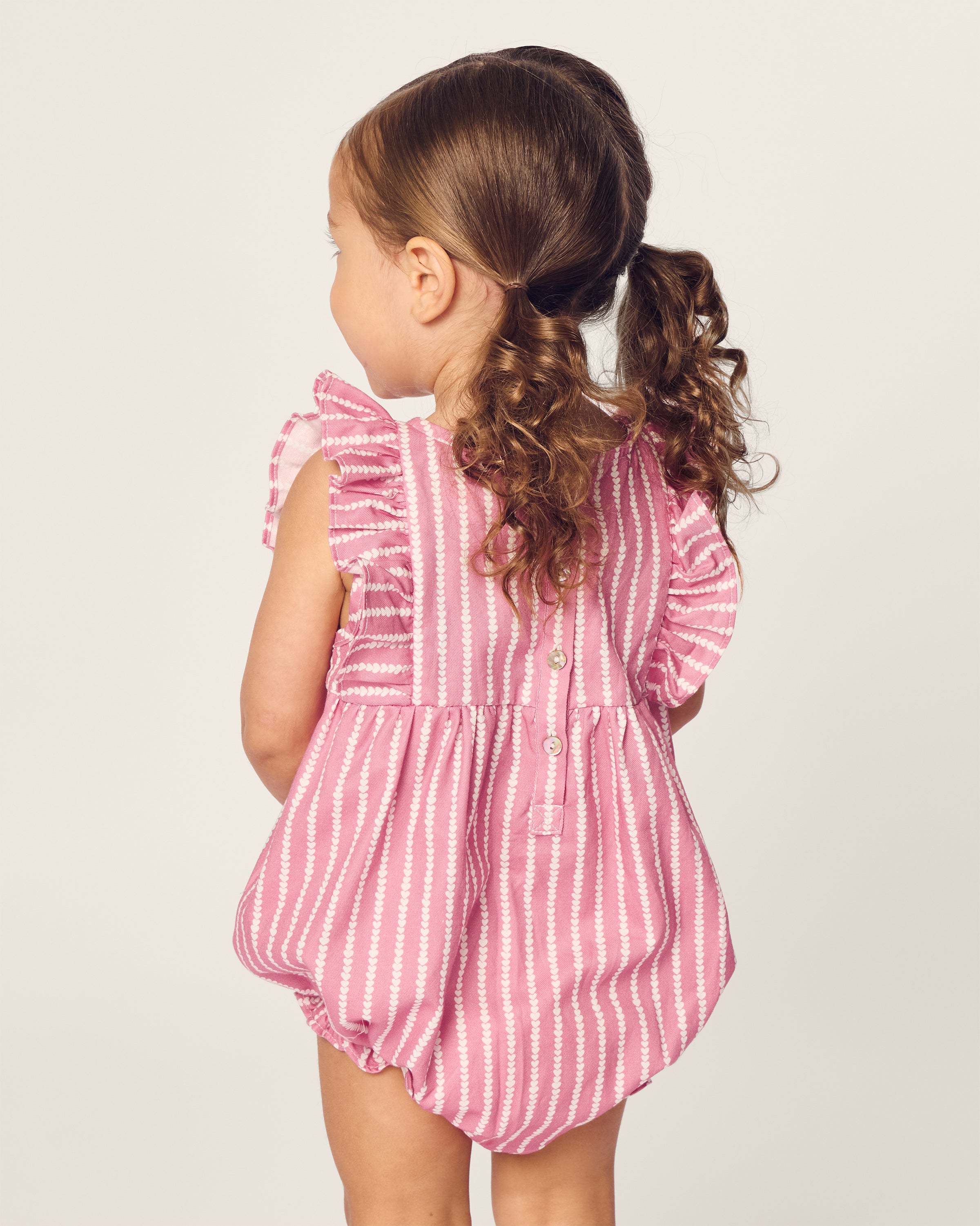 A young child with curly hair, turned away, wears Petite Plumes Babys Twill Ruffled Romper in Love Lines. The pink and white striped Victorian-era design features ruffled sleeves against a plain light-colored background, highlighting its timeless appeal.