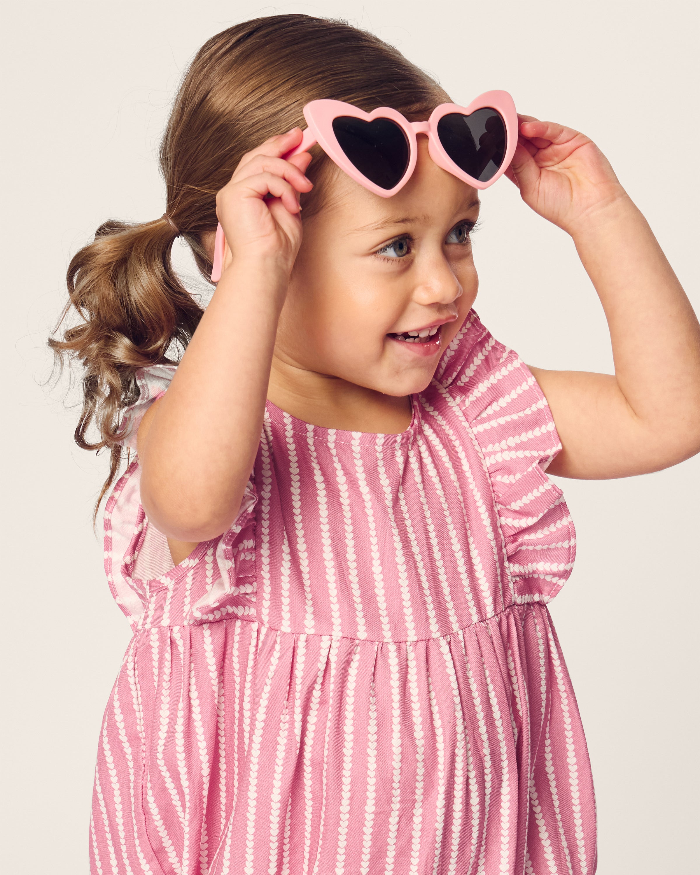 A young girl with light brown pigtails embodies the playful charm of Petite Plumes Babys Twill Ruffled Romper in Love Lines, wearing pink heart-shaped sunglasses and smiling while gazing to the side, dressed in a pink and white striped outfit with ruffled sleeves.