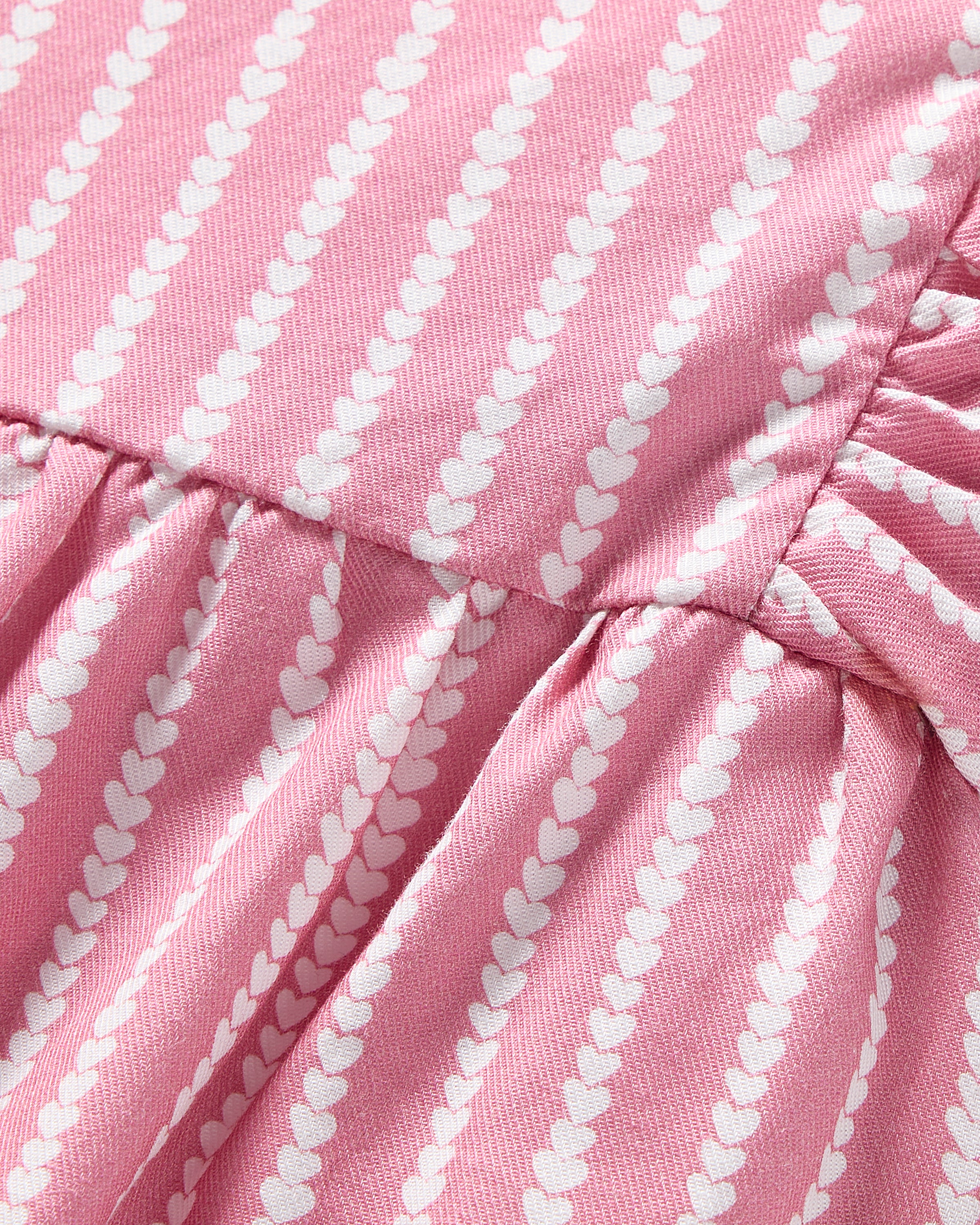 Close-up of Petite Plumes Babys Twill Ruffled Romper in Love Lines, featuring smooth pink fabric with diagonal white heart-shaped lines. A seam gathers slightly, creating a ruffled effect reminiscent of Victorian-era rompers.